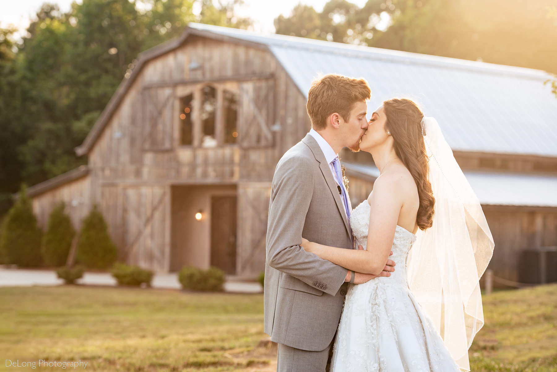 Outdoor portrait of bride and groom kissing during sunset showing the barn in the background at Lady Bird Farms by Charlotte Wedding Photographers DeLong Photography