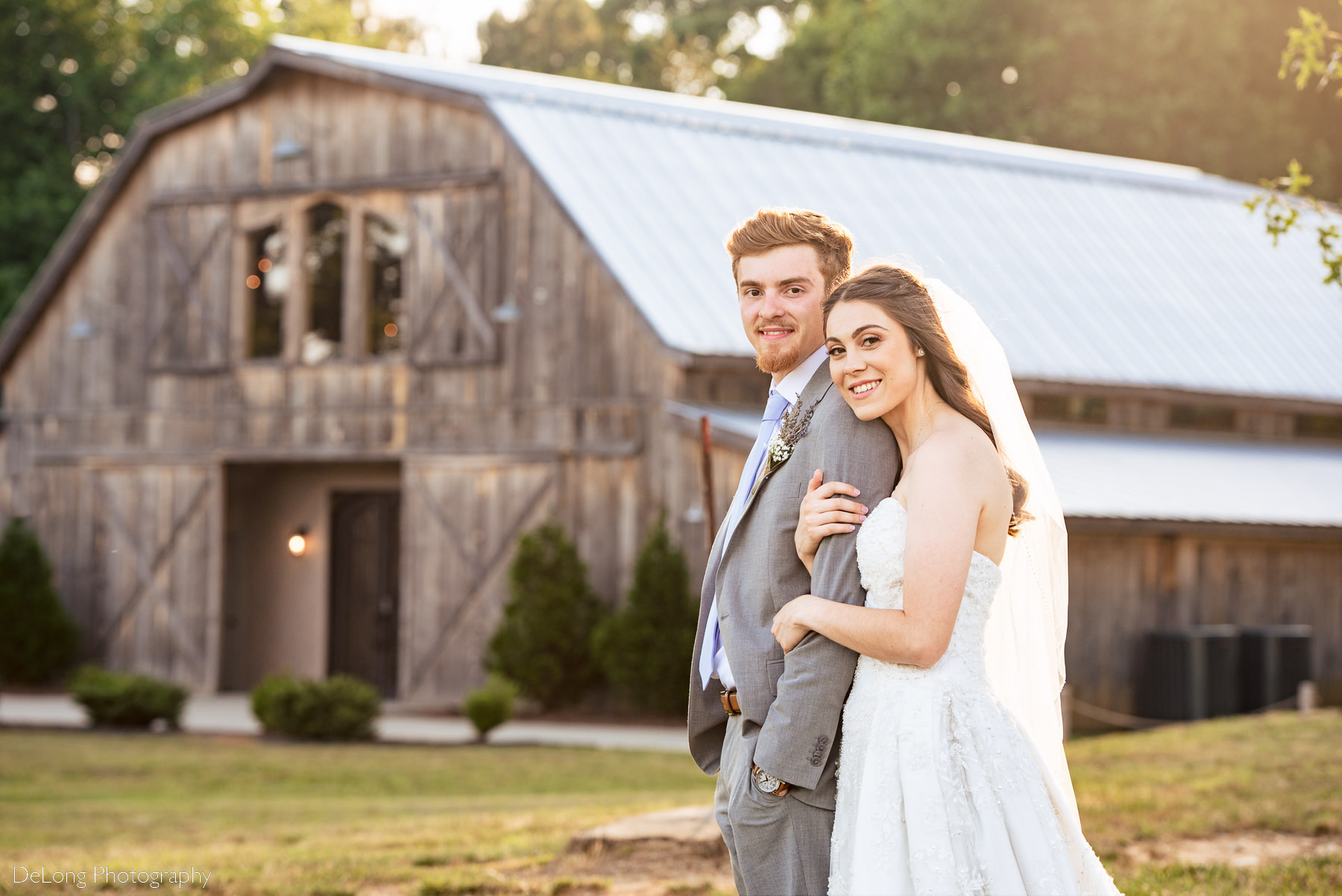 Outdoor portrait of bride and groom snuggling close together during sunset showing the barn in the background at Lady Bird Farms by Charlotte Wedding Photographers DeLong Photography