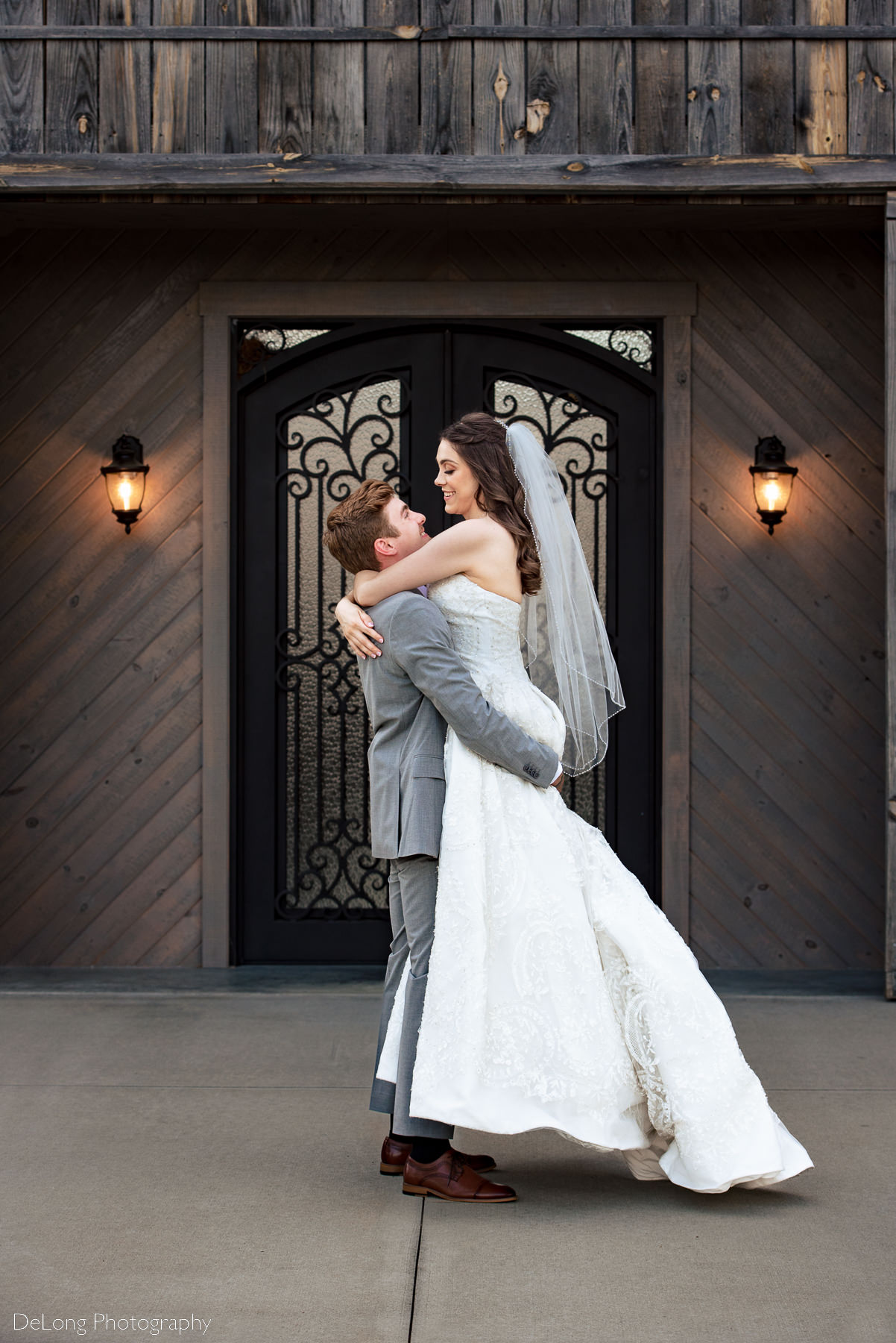 Groom picking his bride up in front of the glass doors of the barn at Lady Bird Farms by Charlotte Wedding Photographers DeLong Photography