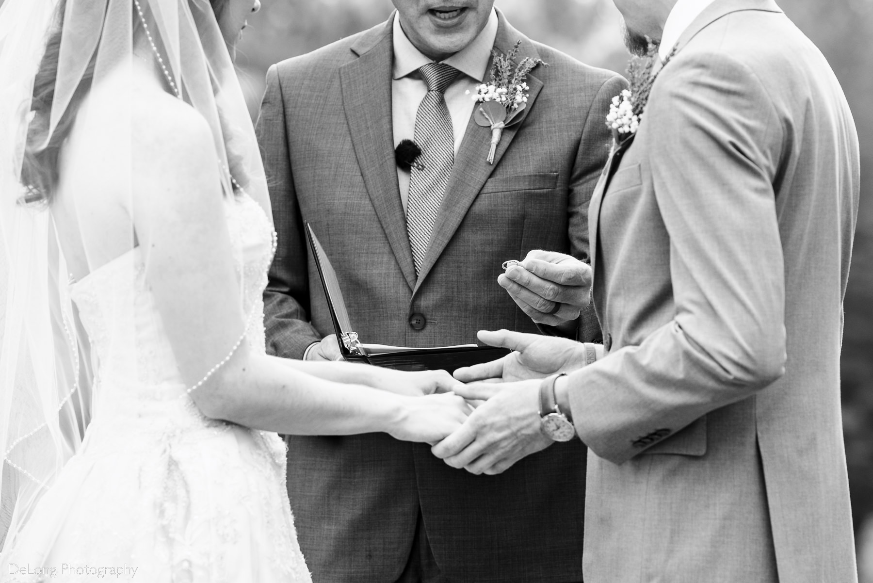 Black and white detail image showing the hands of a bride and groom and an officiant holding a wedding band out during a wedding ceremony by Charlotte Wedding Photographers DeLong Photography