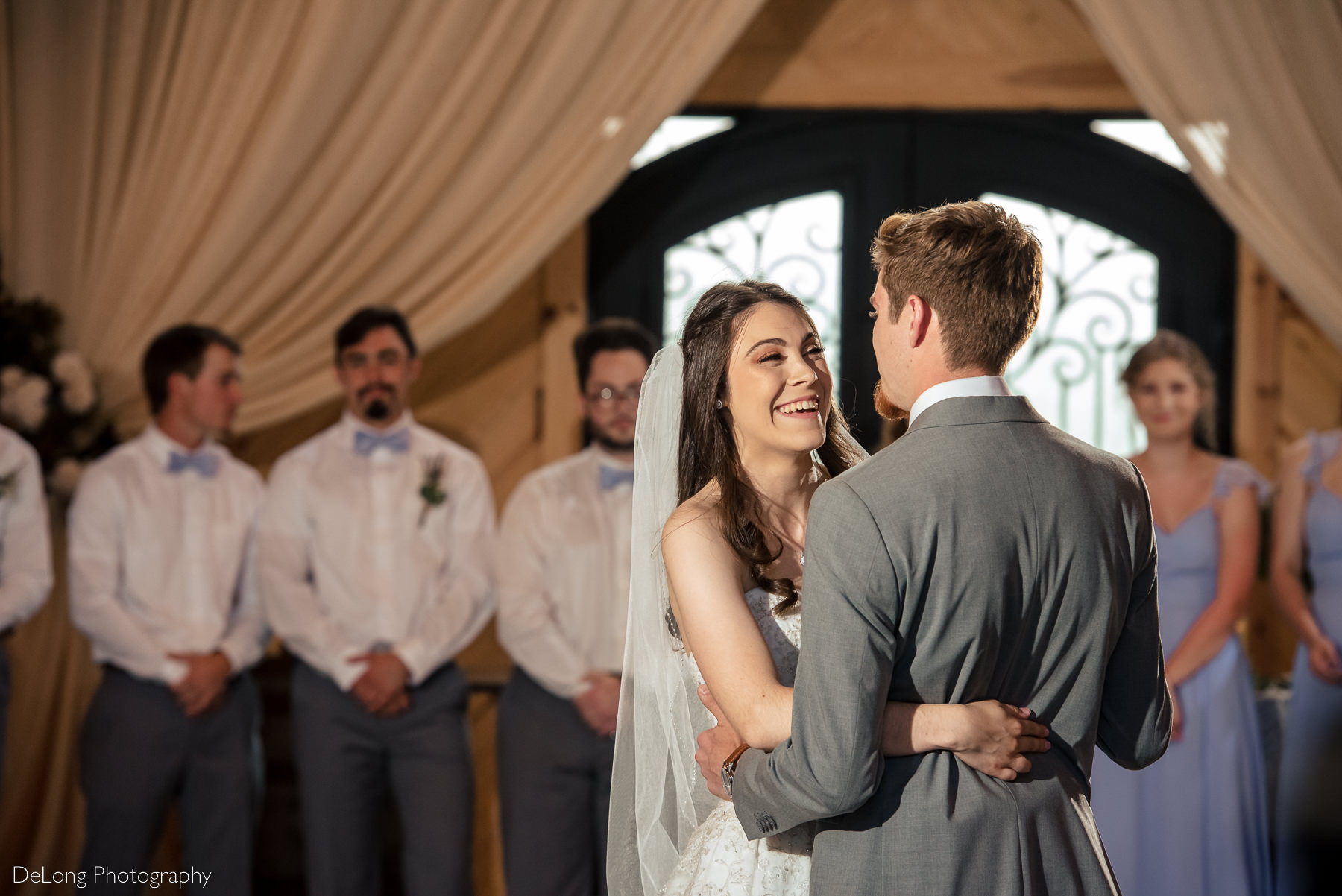 Bride giggling during her first dance with her groom with the wedding party in the background at Lady Bird Farms by Charlotte Wedding Photographers DeLong Photography