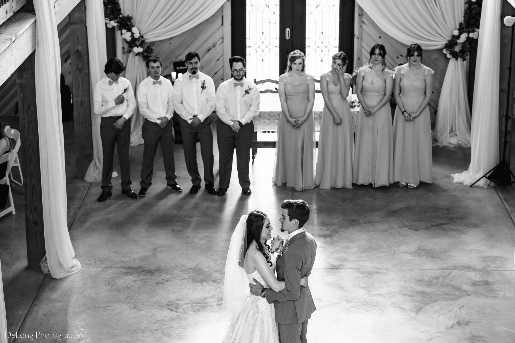 Black and white high perspective photograph of a bride and groom sharing their first dance with their wedding party lined up on the dance floor at Lady Bird Farms by Charlotte Wedding Photographers DeLong Photography