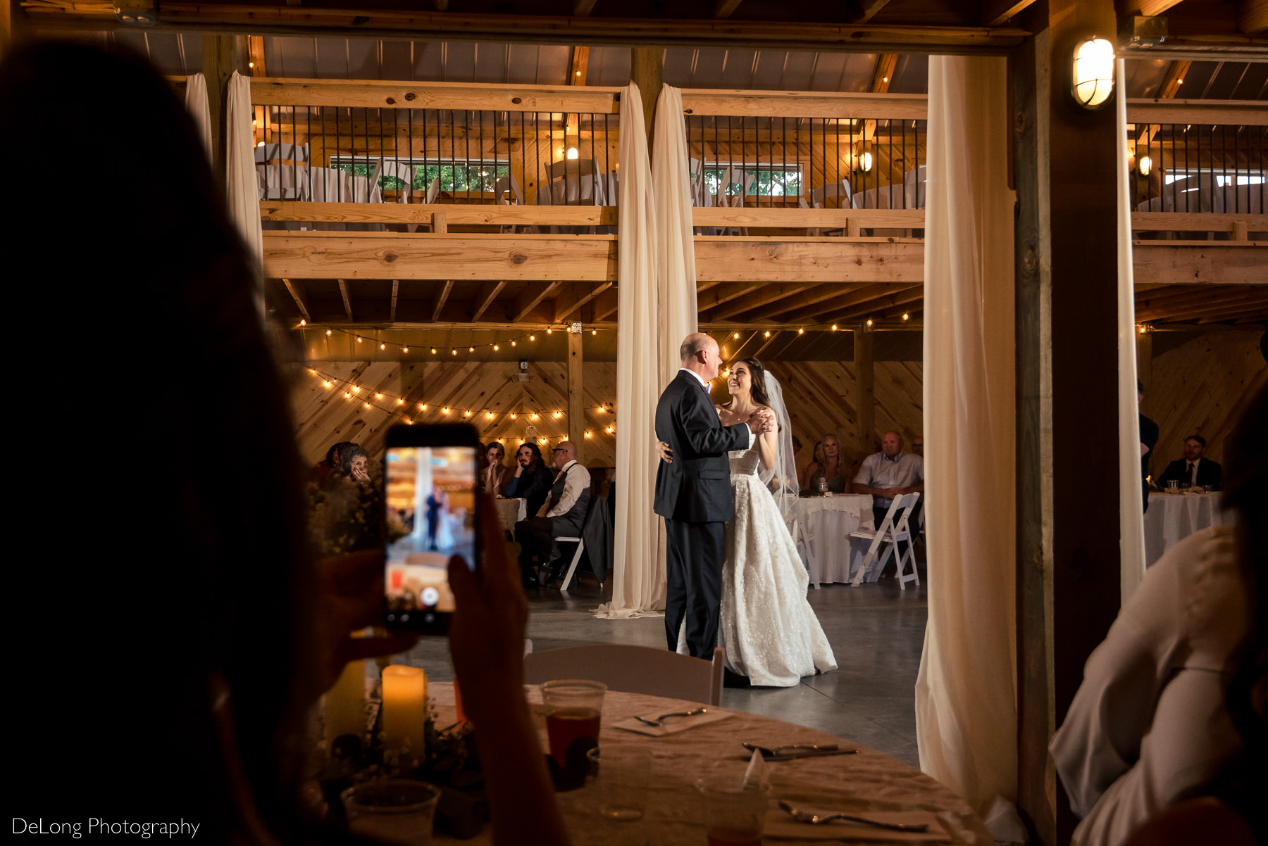 Father-daughter dance image taken from the side of the barn showing the mother of the bride record the dance from her seat at Lady Bird Farms by Charlotte Wedding Photographers DeLong Photography