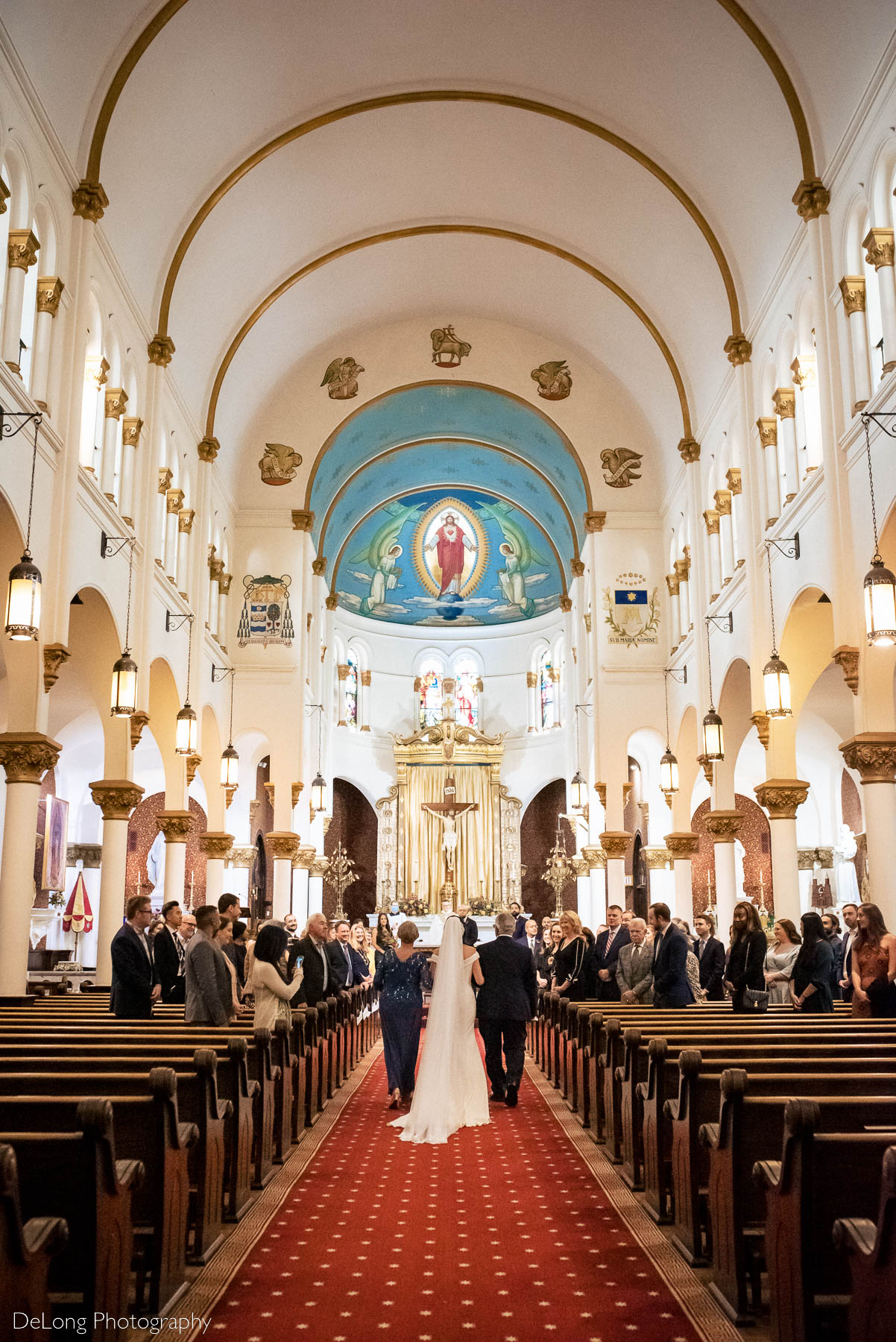 A portrait of a bride being walked down the aisle by her parents showing the expanse and ceiling details inside the church at the Basilica of the Sacred Heart of Jesus in Atlanta, GA by Charlotte wedding photographers DeLong Photography 