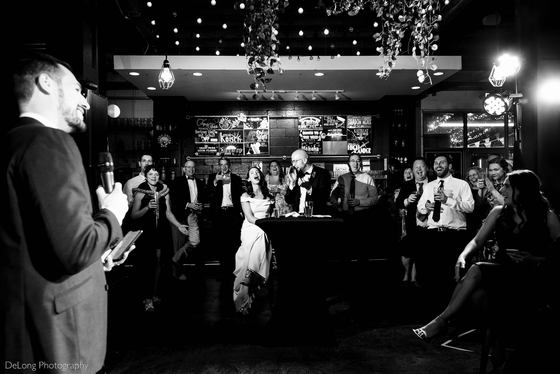 Black and white wide angle photograph of bride and groom and their guests cracking up laughing during a best man speech at Eventide Brewing in Atlanta, GA by Charlotte wedding Photographers DeLong Photography