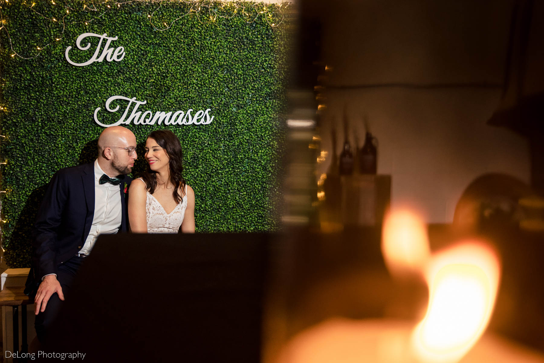 Bride and groom smiling at one another seated under a sign of their last name with candles in the foreground at Eventide Brewing in Atlanta, GA by Charlotte wedding Photographers DeLong Photography