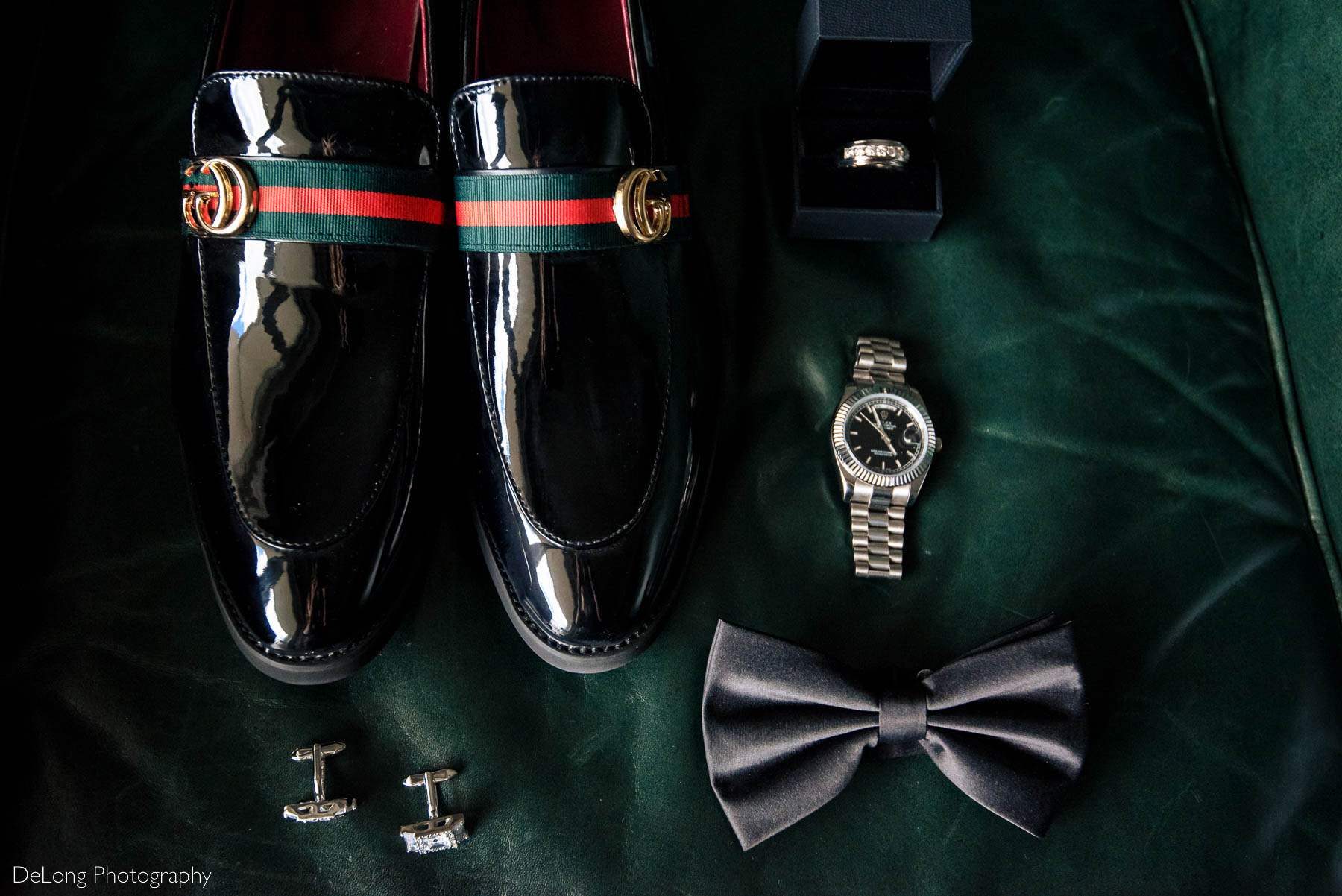 A flat lay of the groom's wedding day details including a pair of black Gucci dress loafers, stainless steel Rolex, diamond cufflinks, and a diamond wedding band on a green leather chair at the Ballantyne Hotel by Charlotte wedding photographers DeLong Photography