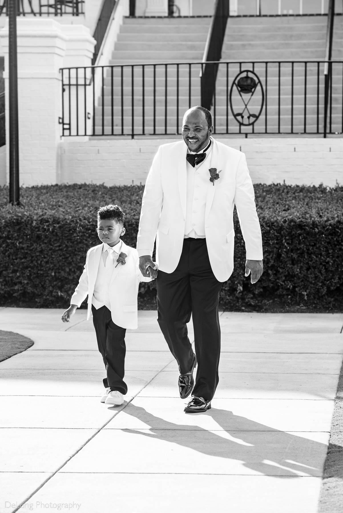 Black and white image of the groom smiling walking down the aisle with one of his twin sons at the beginning of the wedding ceremony at Providence Country Club by Charlotte wedding photographers DeLong Photography