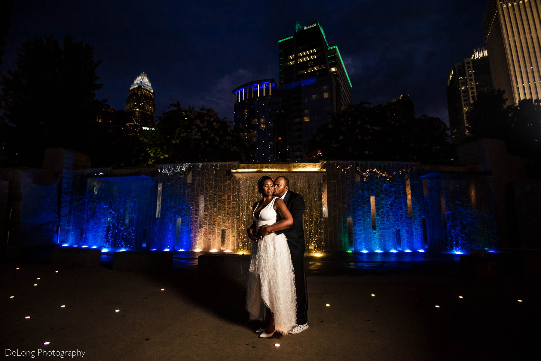 A portrait of the groom giving his bride a kiss on the cheek in front of a blue and yellow water feature at Romare Bearden Park in Uptown Charlotte, NC by Charlotte wedding photographers DeLong Photography