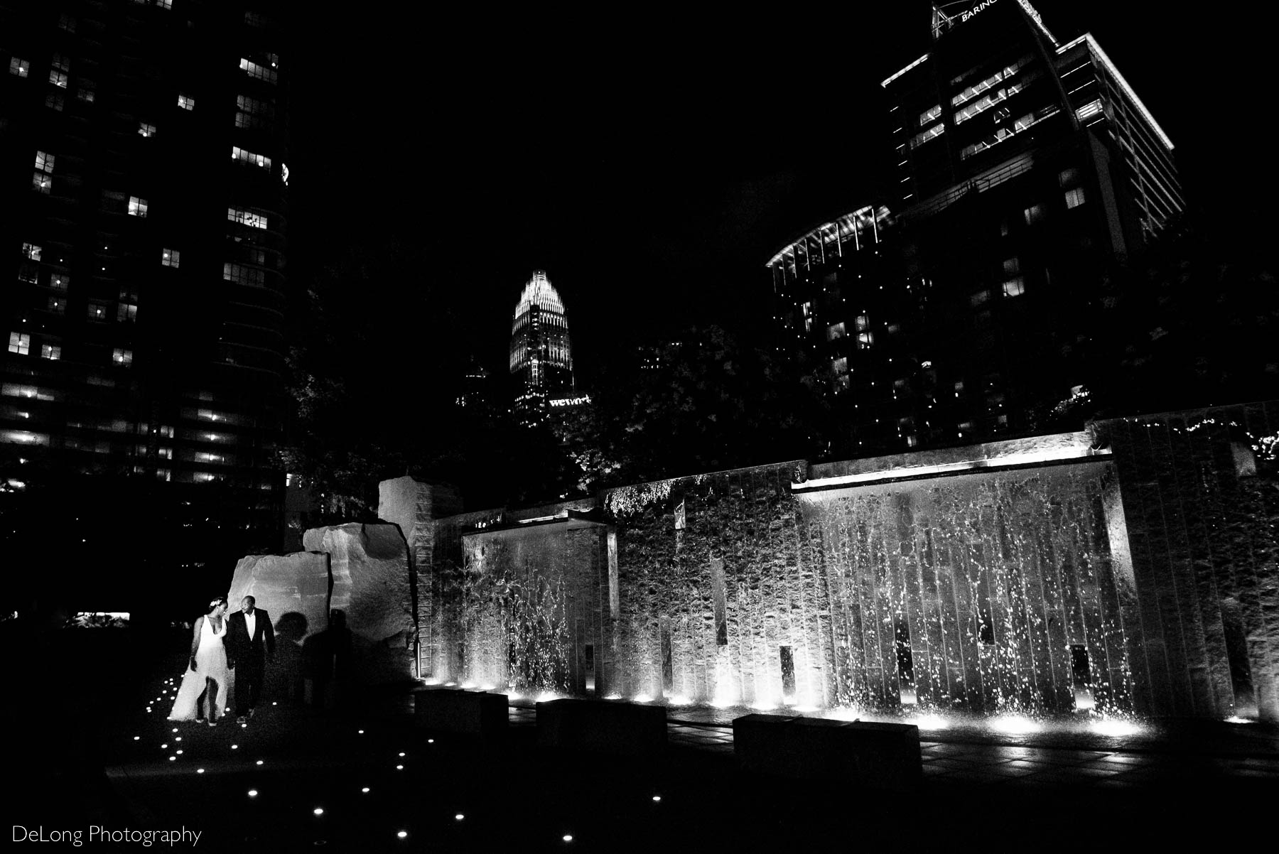 A black and white photograph of the bride and groom small in the frame showcasing the water feature and city skyline at Romare Bearden Park in Uptown Charlotte, NC by Charlotte wedding photographers DeLong Photography