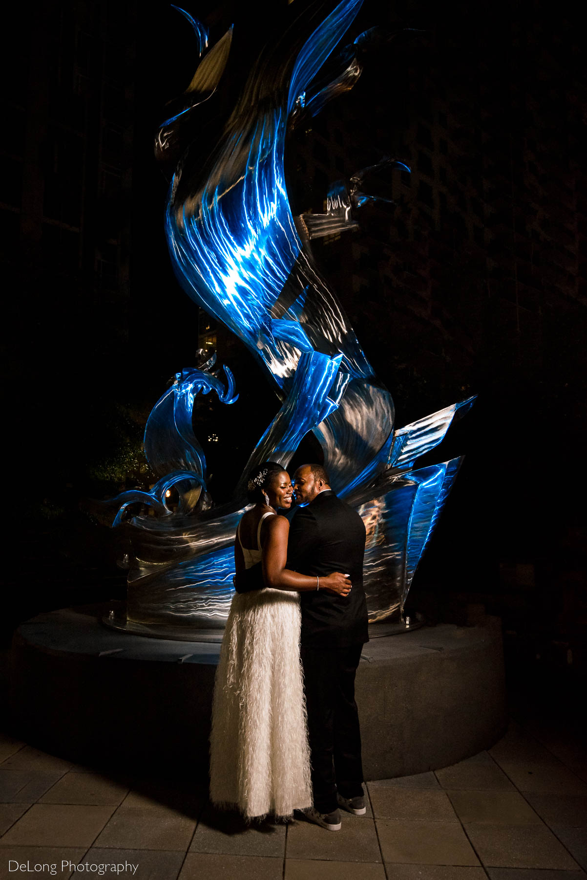 A portrait of a bride and groom where they are mostly turned away from the camera as the groom whispers in the bride's ear making her laugh in front of a blue-lit sculpture at Romare Bearden Park in Uptown Charlotte, NC by Charlotte wedding photographers DeLong Photography