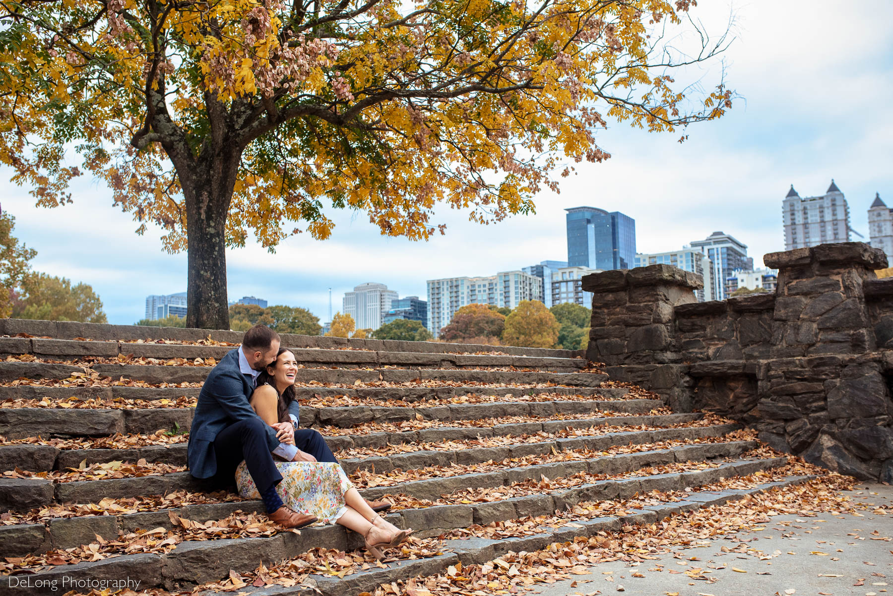 Engaged couple seated on stone steps covered with fall leaves cuddled up and giggling with the city skyline peeking through in the background at Piedmont Park in Atlanta by Charlotte wedding photographers DeLong Photography