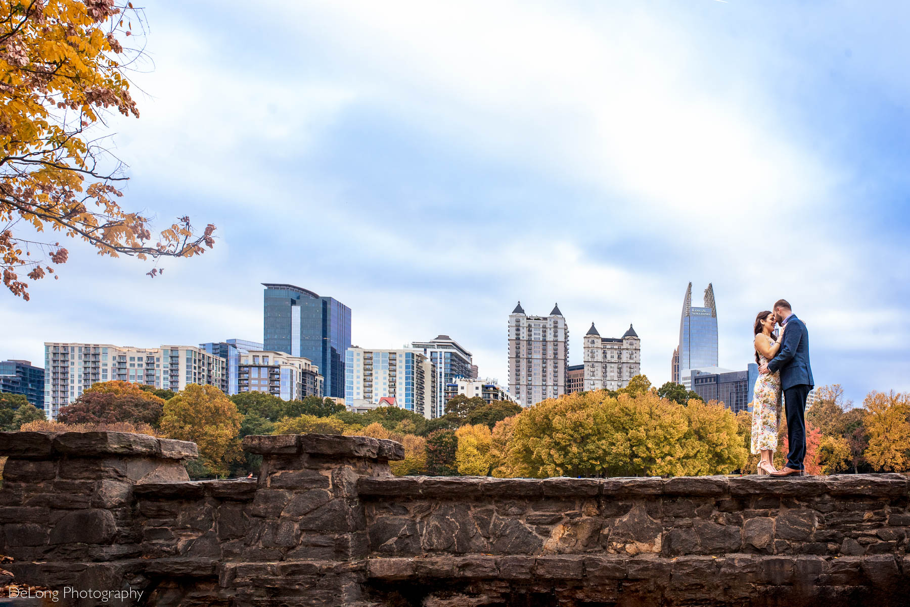 Wide scenery image of the fall foliage and Atlanta city skyline with an engaged couple standing on a stone wall with their foreheads together on the right-hand side at Piedmont Park in Atlanta by Charlotte wedding photographers DeLong Photography