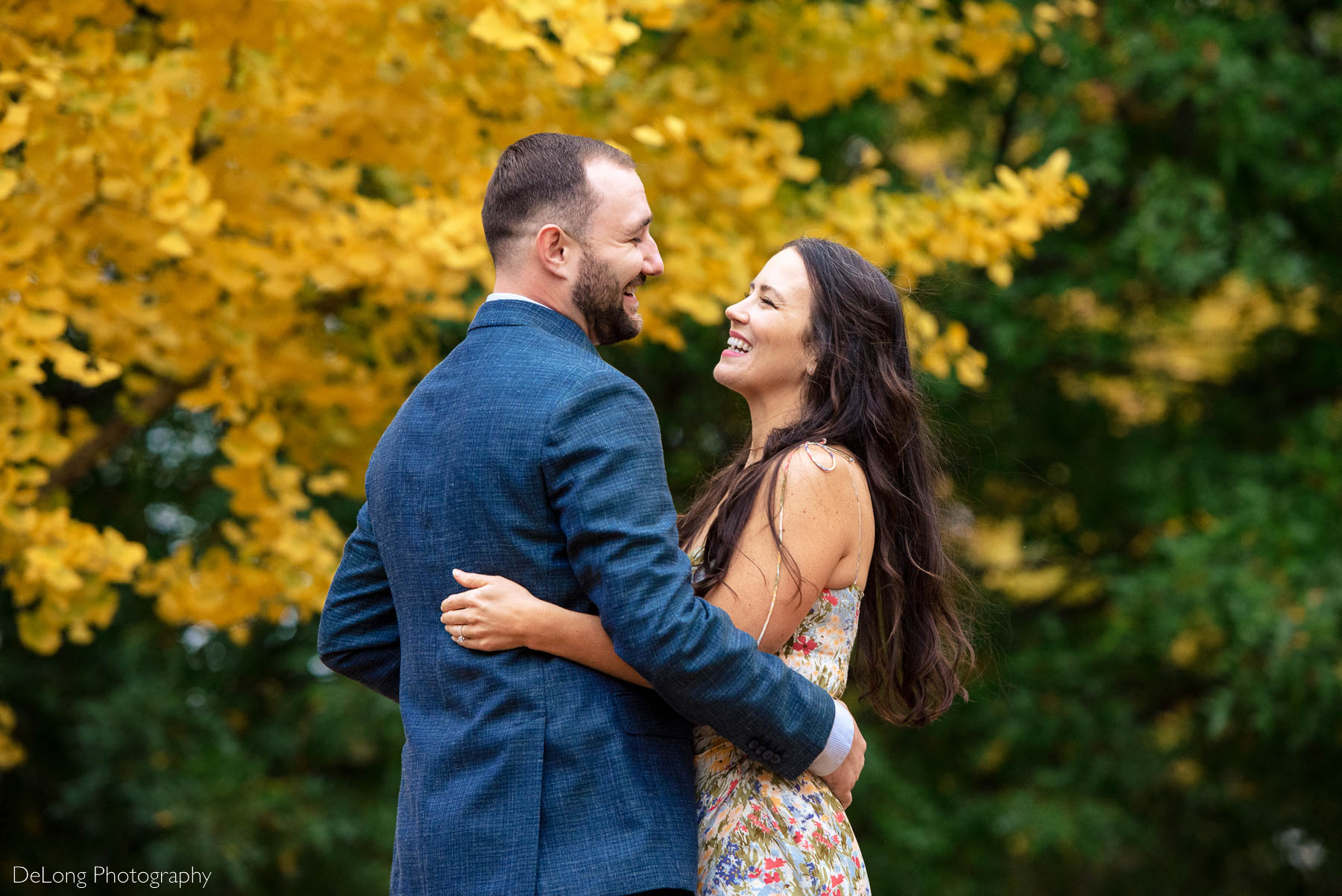 Engaged couple laughing and looking at one another with their arms around each other's waists in front of bright yellow and deep green fall foliage at Piedmont Park in Atlanta by Charlotte wedding photographers DeLong Photography