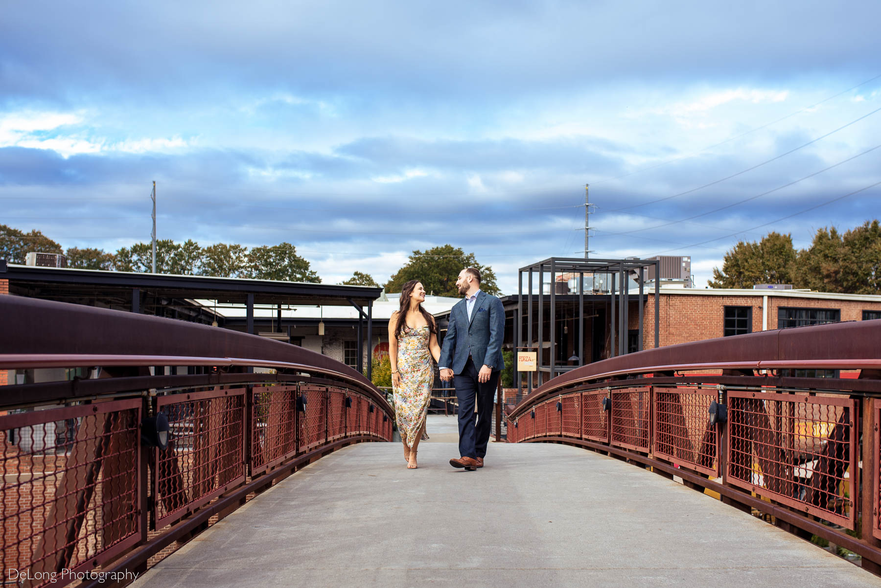 Engaged couple smiling at one another walking toward the camer on the bridge at Westside Provisions in Atlanta by Charlotte Wedding Photographers DeLong Photography