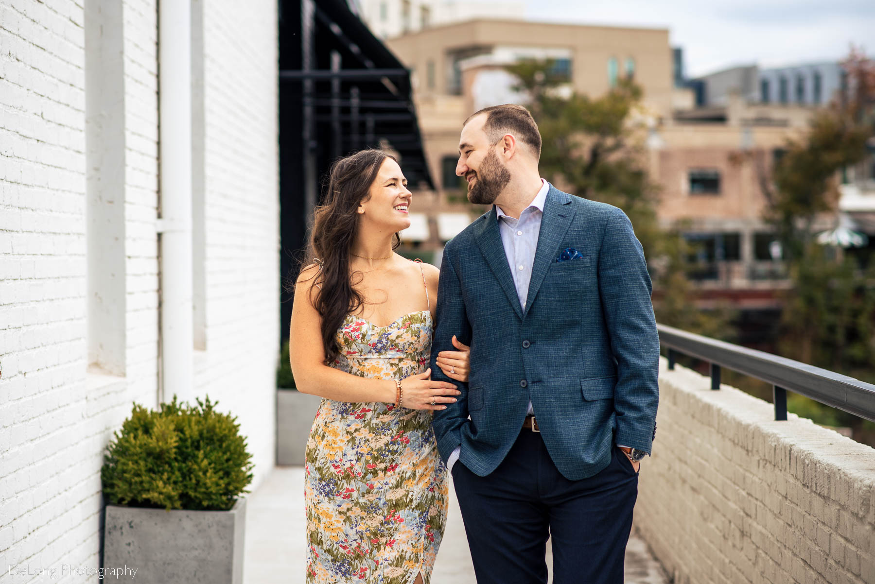 Medium crop image of an engaged couple smiling at one another walking arm-in-arm on a sidewalk next to a building that has white brick walls  and modern square concrete planters at Westside Provisions in Atlanta by Charlotte Wedding Photographers DeLong Photography