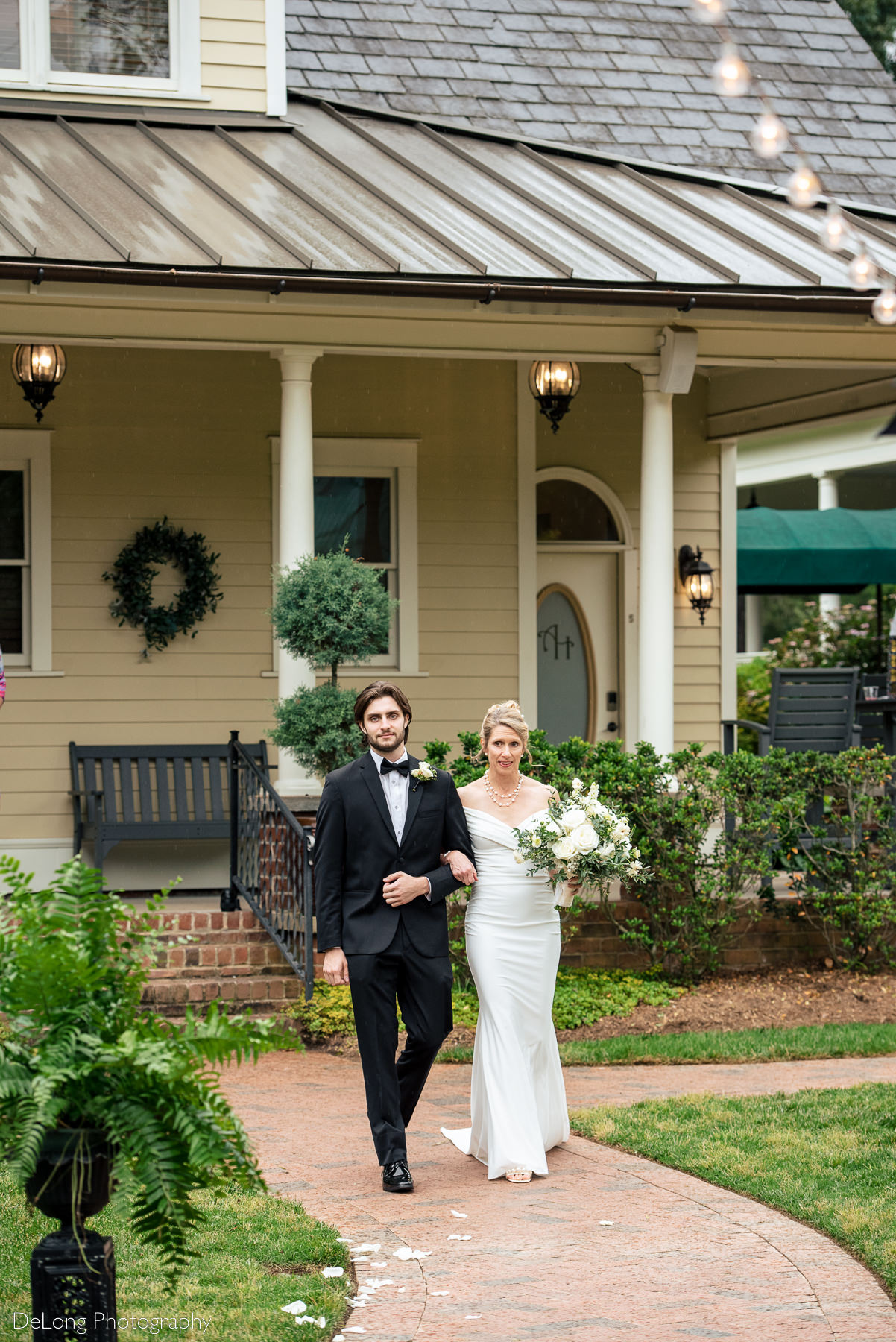 Bride being walked down the aisle by her teenage son during an outdoor wedding ceremony at Alexander Homestead. Photograph by Charlotte wedding photographers DeLong Photography.