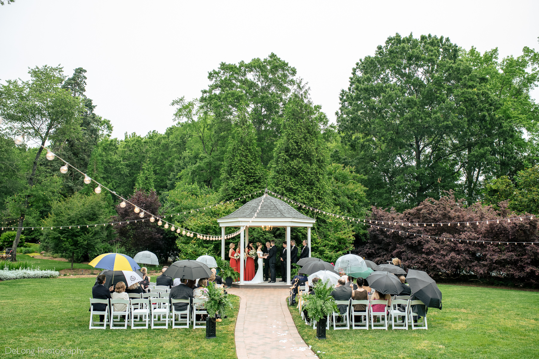 Wide photograph taken from a distance of a wedding ceremony at Alexander Homestead on a rainy day. Photograph by Charlotte wedding photographers DeLong Photography.