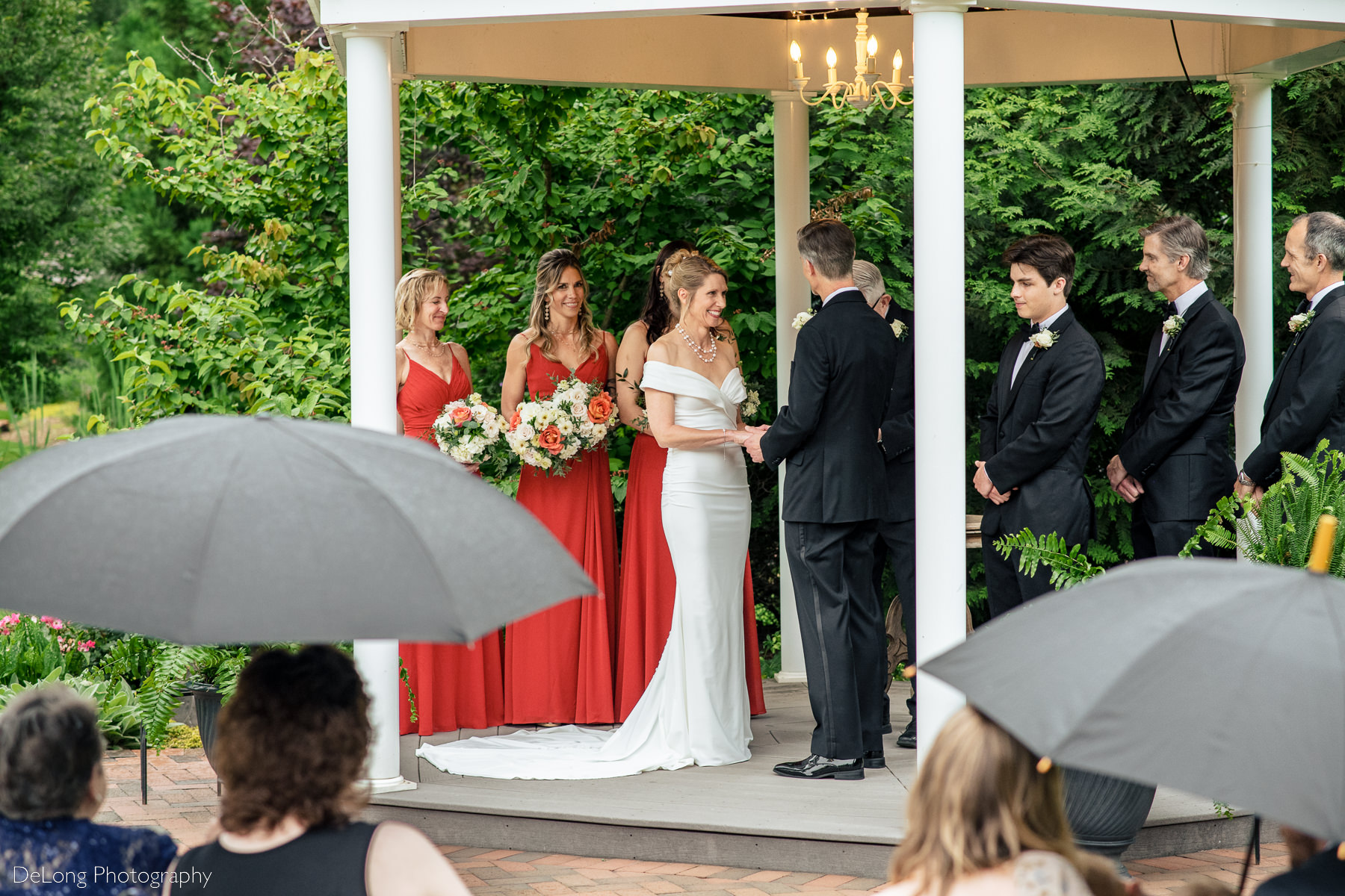 Bride smiling at her groom during a wedding ceremony at Alexander Homestead. She is framed by umbrellas as it was rainy. Photograph by Charlotte wedding photographers DeLong Photography.