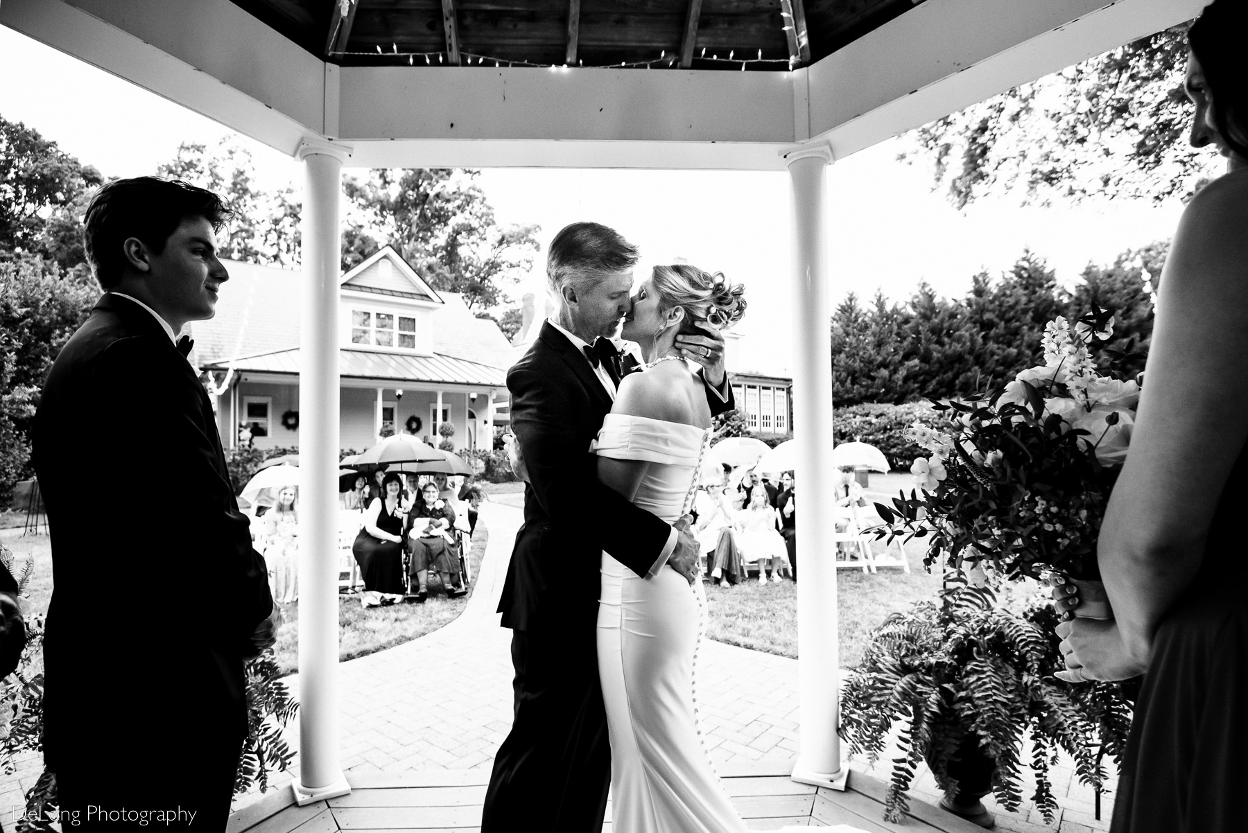 Black and white photograph taken from behind the bride and groom during their first kiss at Alexander Homestead. Photograph by Charlotte wedding photographers DeLong Photography.