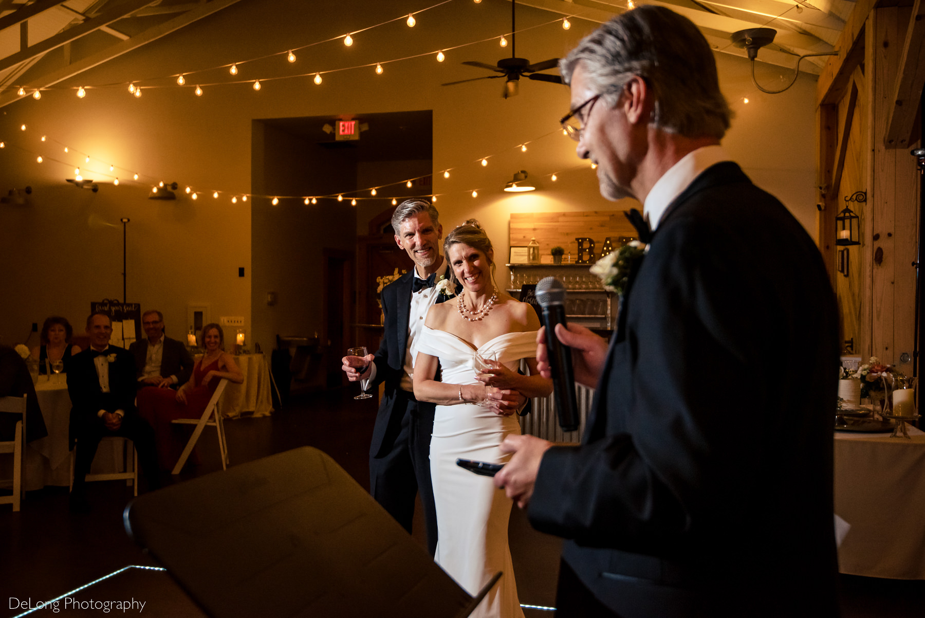 Bride and groom smiling at the best man during his toast at Alexander Homestead. Photograph by Charlotte wedding photographers DeLong Photography.