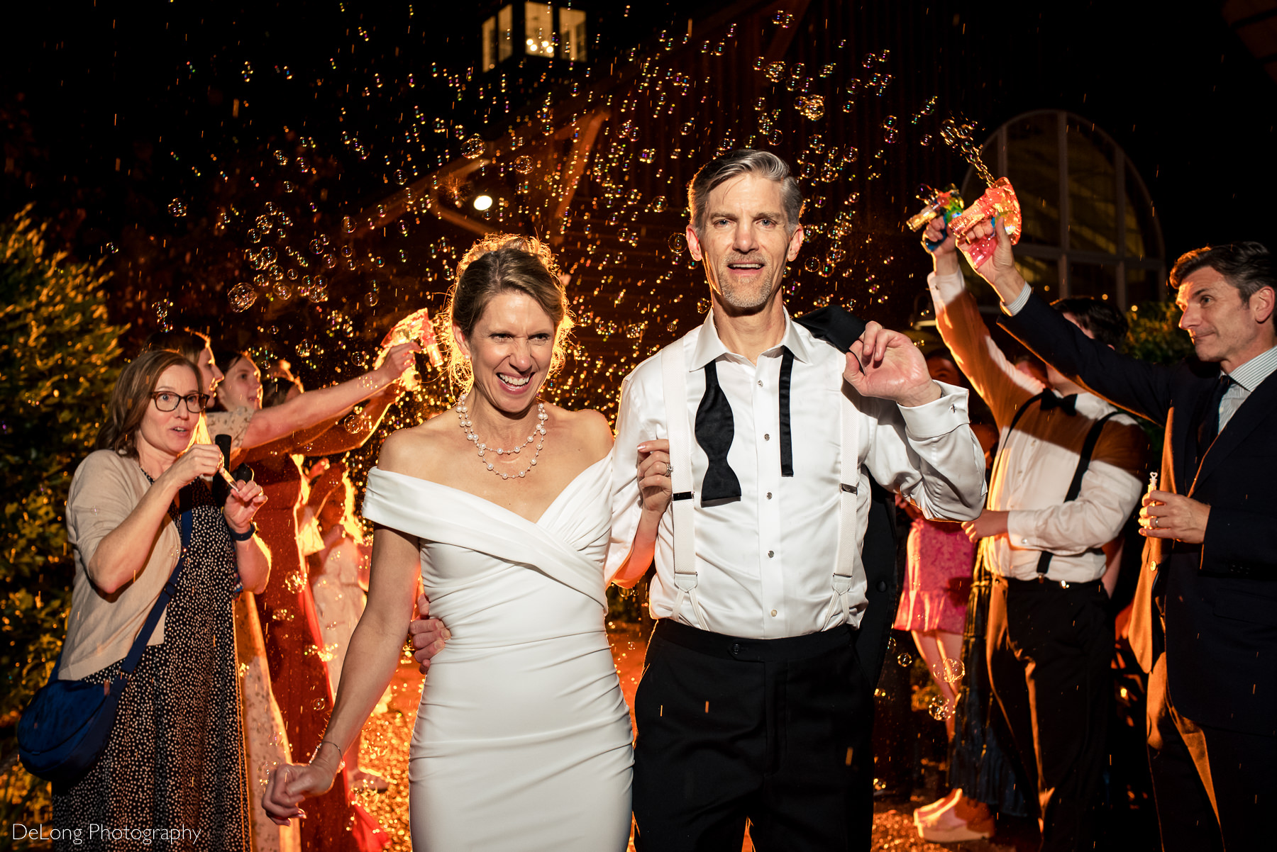 Candid photograph of bride and groom exiting their wedding reception surrounded by bubbles and an orange glow of light behind them. 