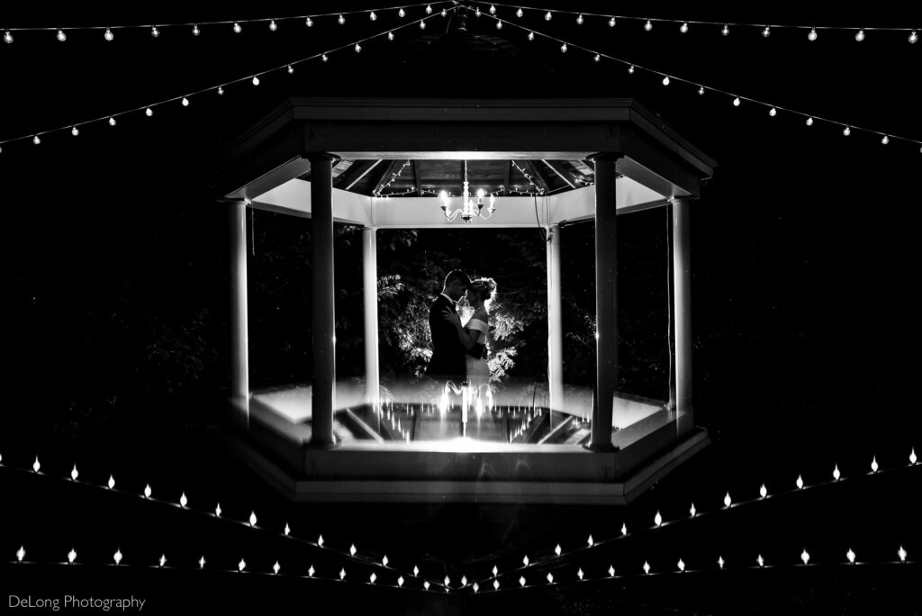 Black and white nighttime silhouette of a couple with their foreheads together, standing inside the gazebo at Alexander Homestead. Photo by Charlotte wedding photographers DeLong Photography.