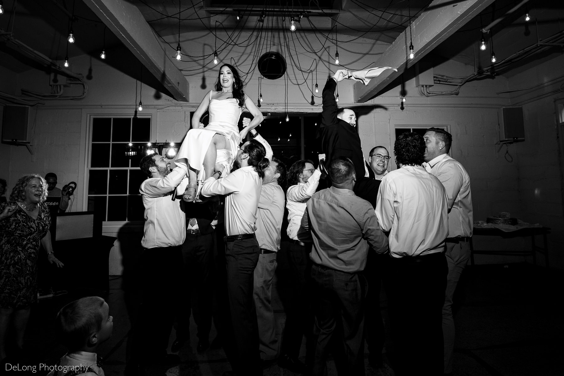 Black and white candid photograph of  the bride and groom being lifted in the air on chairs by their guests during their wedding reception at Upstairs Atlanta. Photograph by Charlotte wedding photographers DeLong Photography.