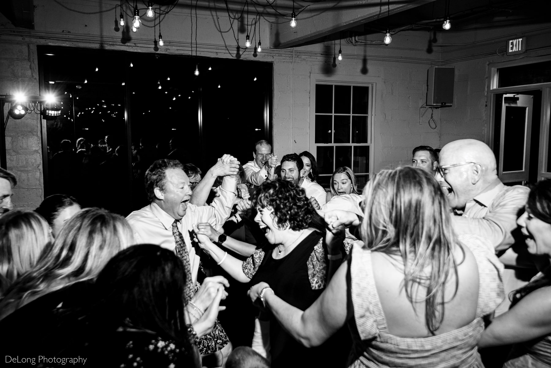 Black and white photograph of guests reacting in excitement and exhilaration as they race toward the center of the dance floor from all sides while holding hands. Photograph by Charlotte wedding photographers DeLong Photography.