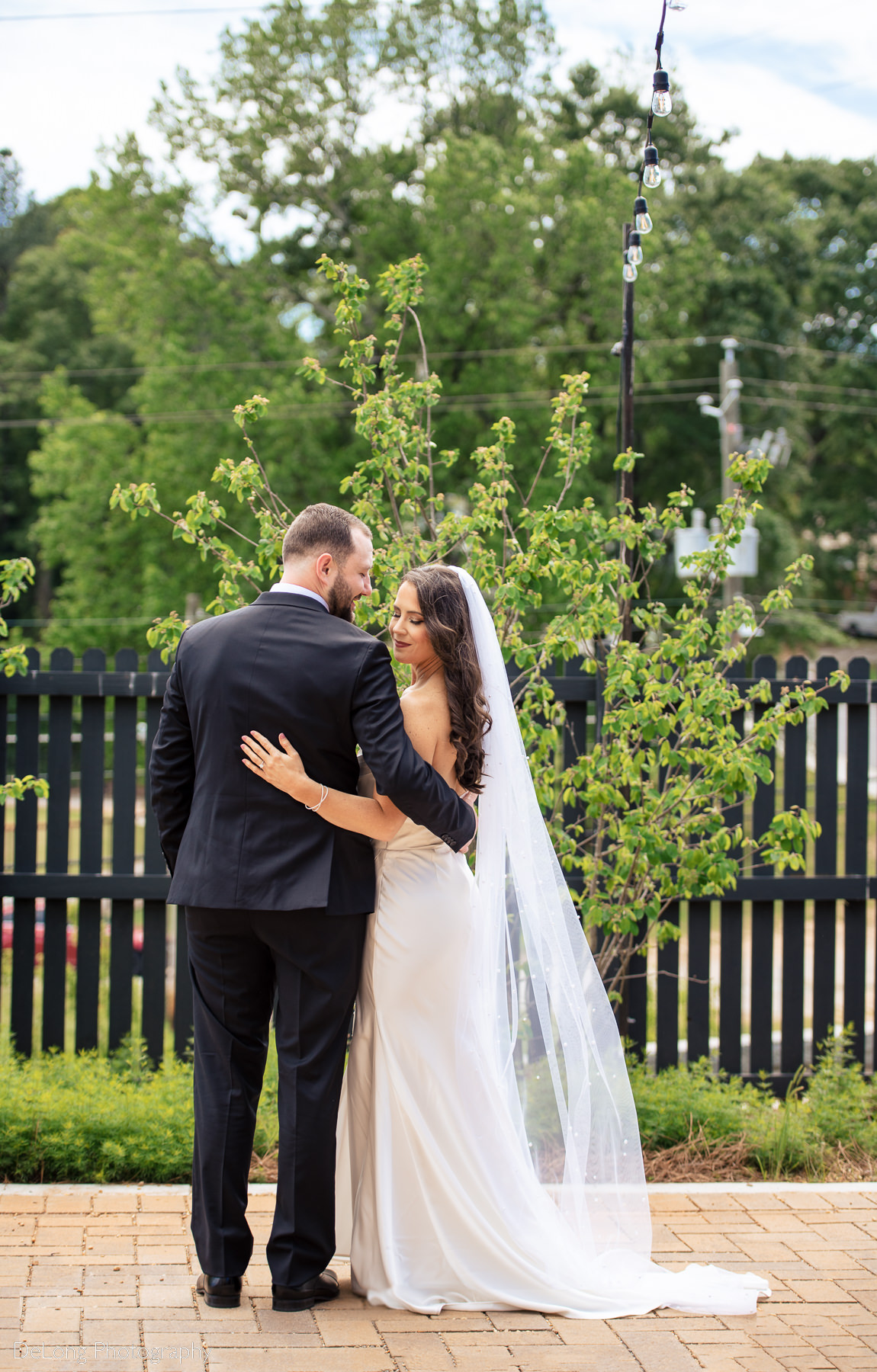 Bride and groom with their arms around each other, facing away from the camera. The bride looks back and down toward her train, softly smiling, while the groom gazes at her lovingly. Photo taken outside Upstairs Atlanta by Charlotte wedding photographers, DeLong Photography.