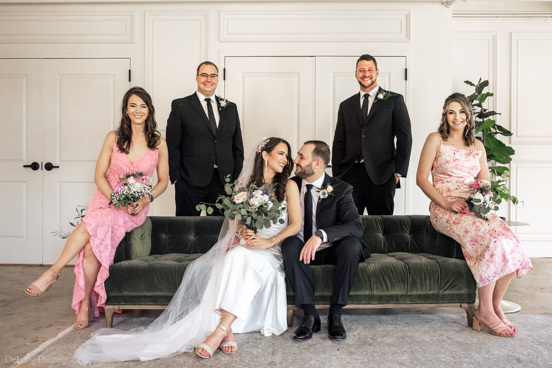 Bride and groom smiling at one another snuggled on a green velvet sofa surrounded by their wedding party. Photograph by Charlotte wedding photographers DeLong Photography.