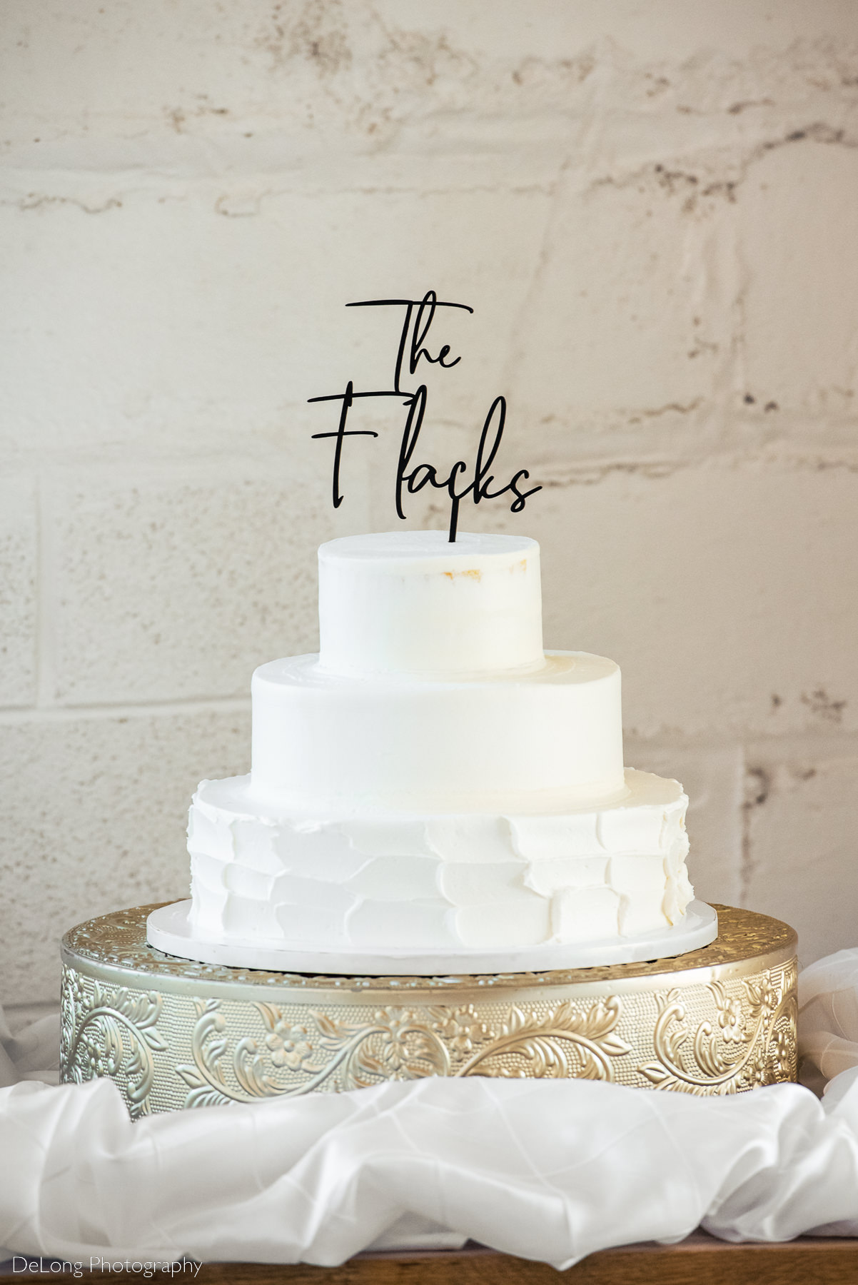 Photograph of a simple and modern wedding cake at Upstairs Atlanta with a black cake topper reading the bride and groom's last name in a handwritten front. Photograph by Charlotte wedding photographers DeLong Photography.