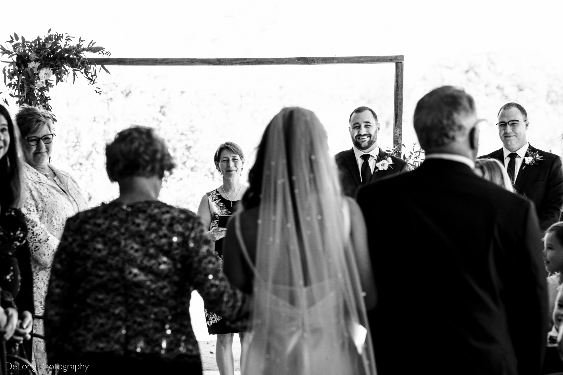 Black and white photograph focused on a smiling groom taken behind the bride as she is walked down the aisle by her parents. Photograph by Charlotte wedding photographers DeLong Photography.