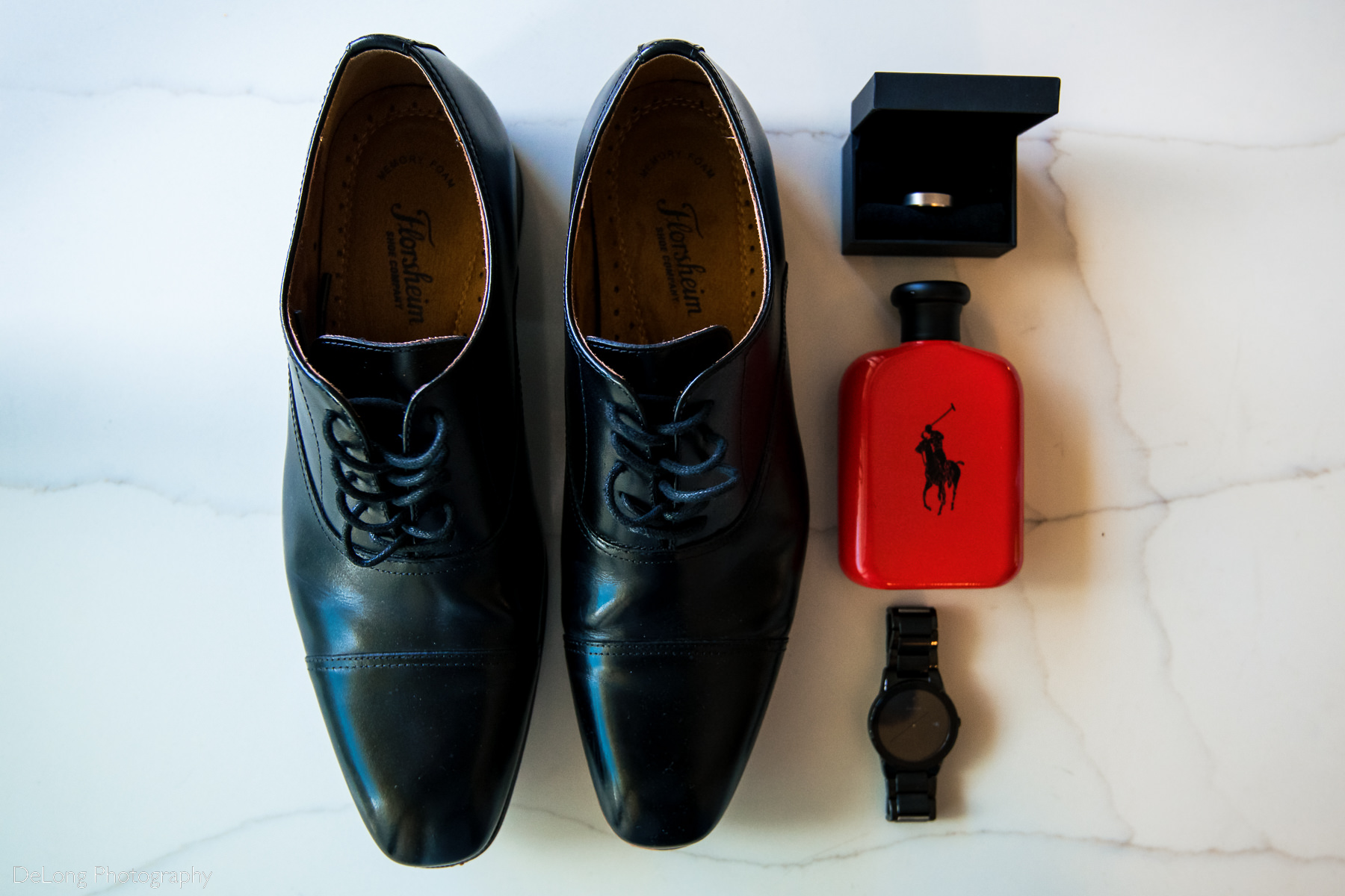 Photograph of groom's black details, including dress shoes his watch and ring box, alongside a pop of red Polo cologne on a marble background by Charlotte wedding photographers DeLong Photography