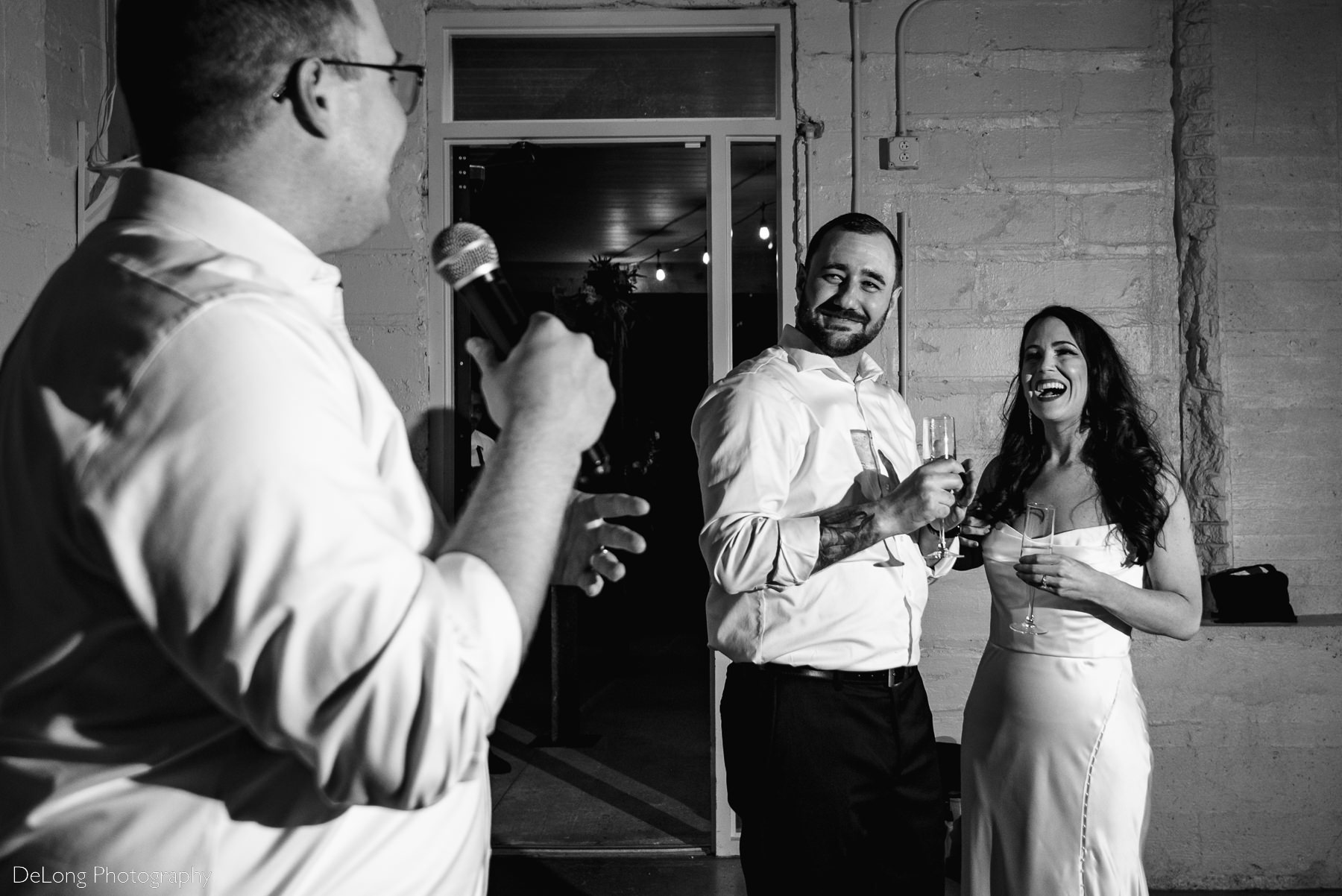 Black and white photograph of a bride and groom smiling in response to the best man's speech at Upstairs Atlanta. Photograph by Charlotte wedding photographers DeLong Photography.