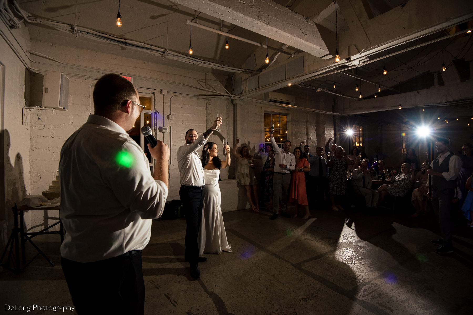 Wide angle photograph taken over the best man's shoulder showing the bridge and groom raising a glass to his speech. Photograph by Charlotte wedding photographers DeLong Photography.