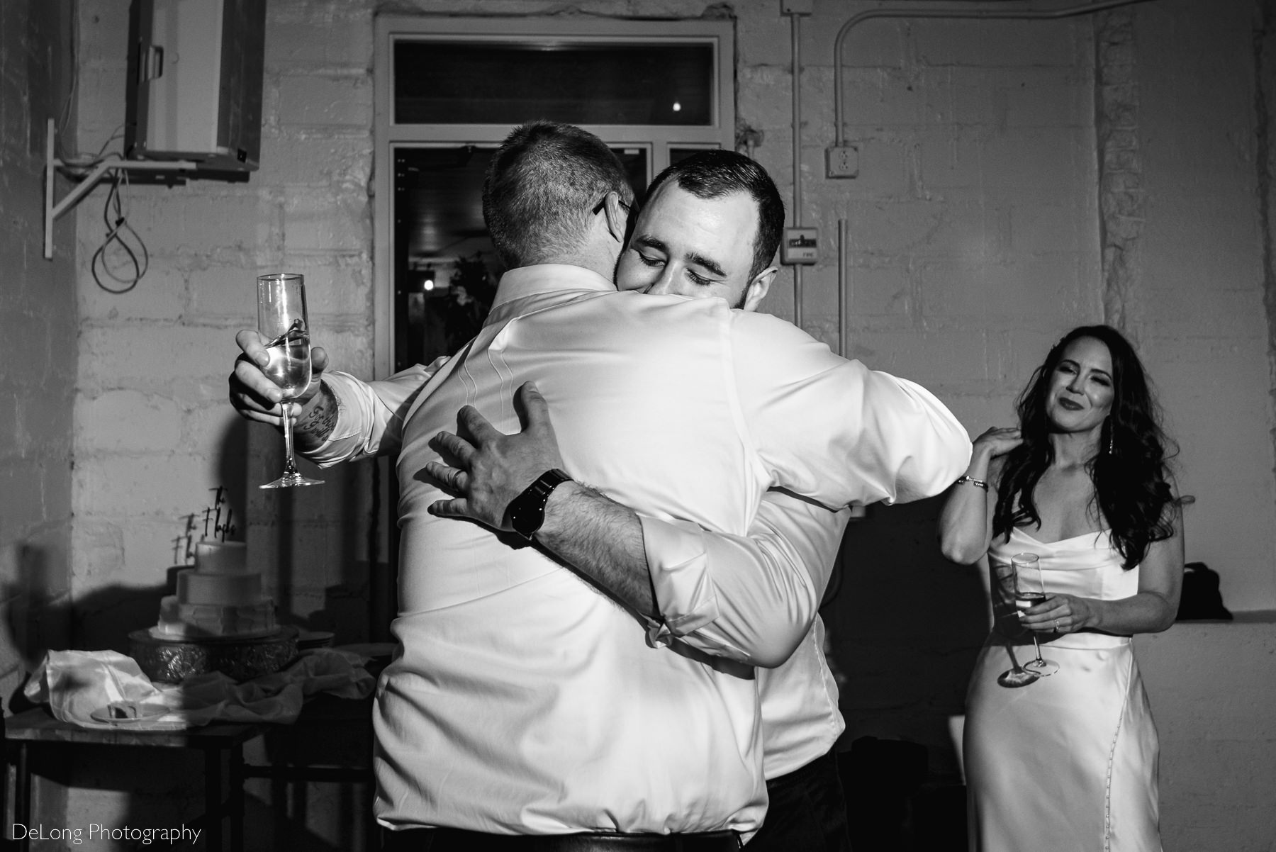 Black and white photograph of the groom tightly hugging the best man as his bride looks on lovingly. Photograph by Charlotte wedding photographers DeLong Photography.