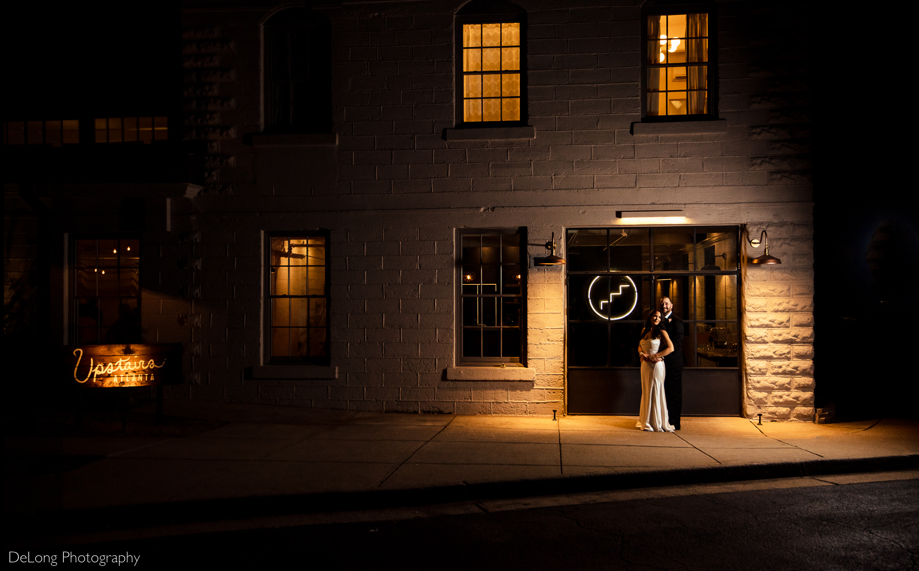 Wide angle nighttime portrait of a bride and groom outside Upstairs Atlanta in Atlanta, Georgia. They are surrounded by the orange glow of the outdoor lamps. The Upstairs Atlanta sign is lit up and included in the photograph. Photograph by Charlotte wedding photographers DeLong Photography.