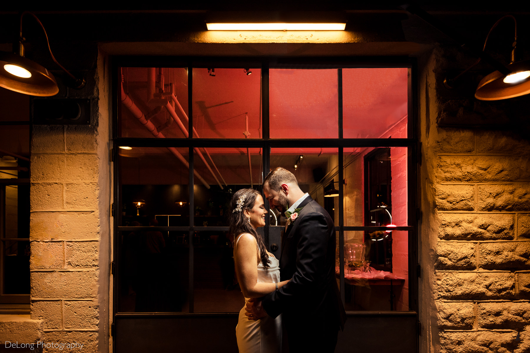 Bridge and groom smiling with their foreheads together outside Upstairs Atlanta at night. Their are surrounded by the orange glow of the outdoor lamps. A pink light illuminates the inside of the building. Photograph by Charlotte wedding photographers DeLong Photography.