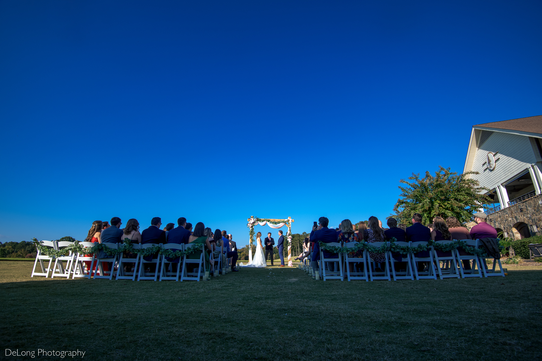 Wide low-angle photograph showcasing the set up during a wedding ceremony on the lawn of Pine Island Country Club with a cloudless blue sky by Charlotte wedding photographers DeLong Photography
