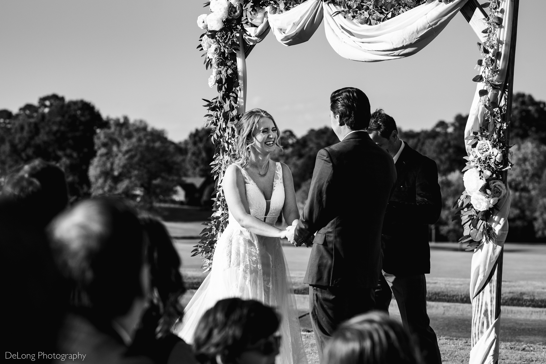 Black and white image of a bride cracking up laughing while holding hands with her groom during their wedding ceremony at Pine Island Country Club by Charlotte wedding photographers DeLong Photography