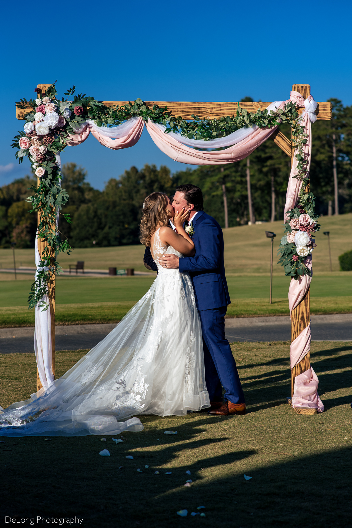 Bride and groom sharing their first kiss under a blush, burgundy, and white floral decorated arbor at Pine Island Country Club by Charlotte wedding photographers DeLong Photography