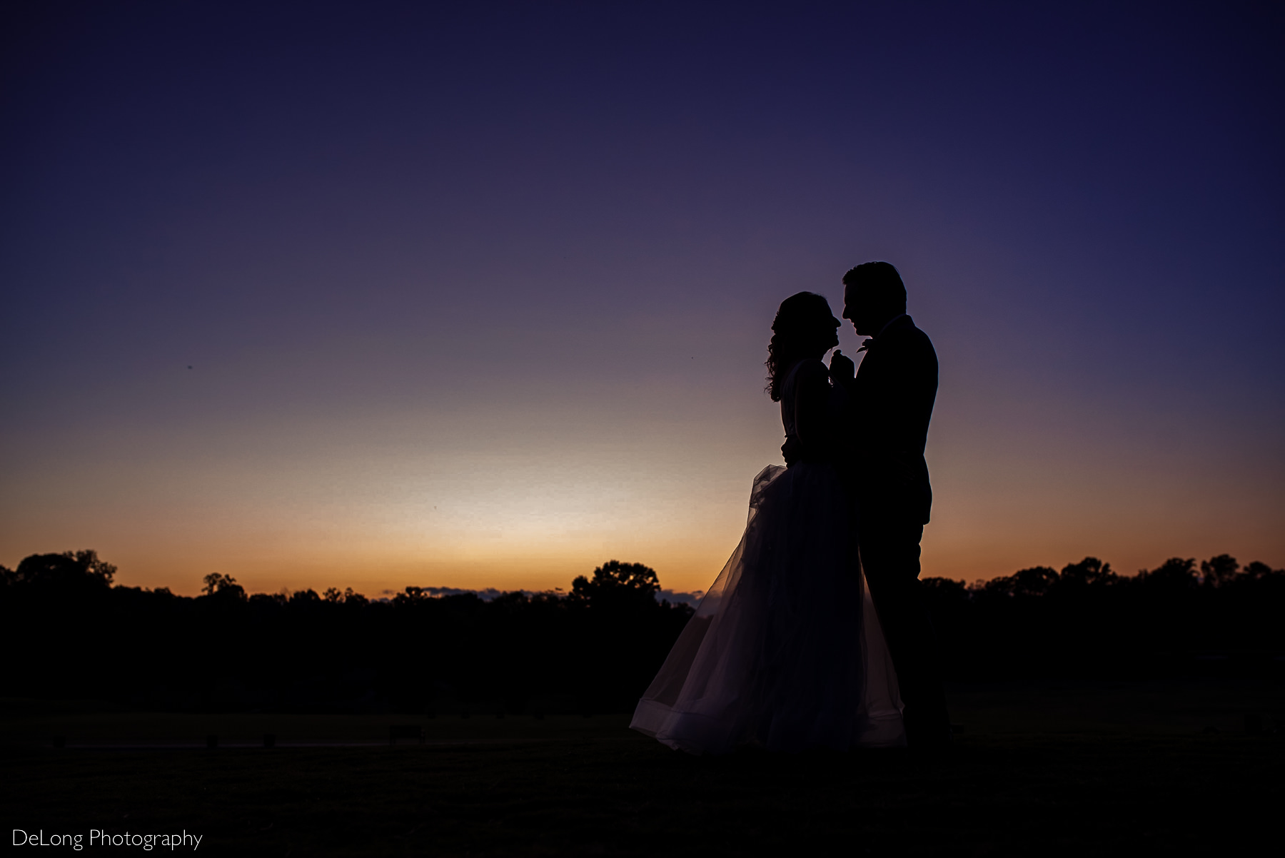 Purple and orange sunset photograph of a bride and groom smiling just before he lifts her chin for a kiss at Pine Island Country Club in Charlotte, NC by Charlotte wedding Photographers DeLong Photography
