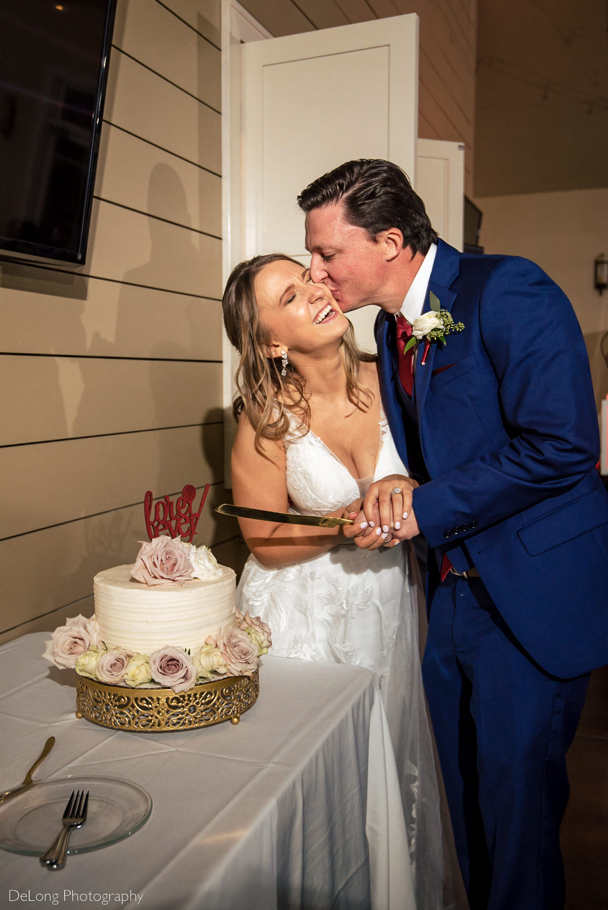 Groom kissing bride on the cheek who is laughing while they hold their cake knife before cutting their wedding cake at Pine Island Country Club in Charlotte, NC by Charlotte wedding Photographers DeLong Photography