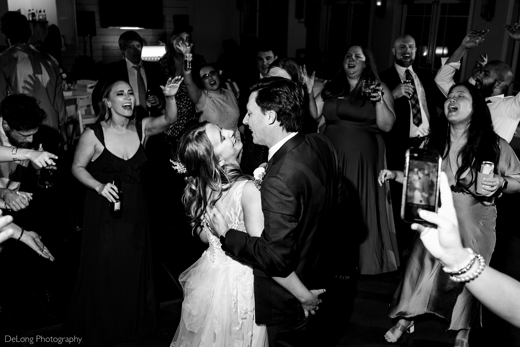 Black and white image of bride and groom with arms around each other singing along with all of their friends who encircled them on the dance floor in Charlotte, NC by Charlotte wedding Photographers DeLong Photography