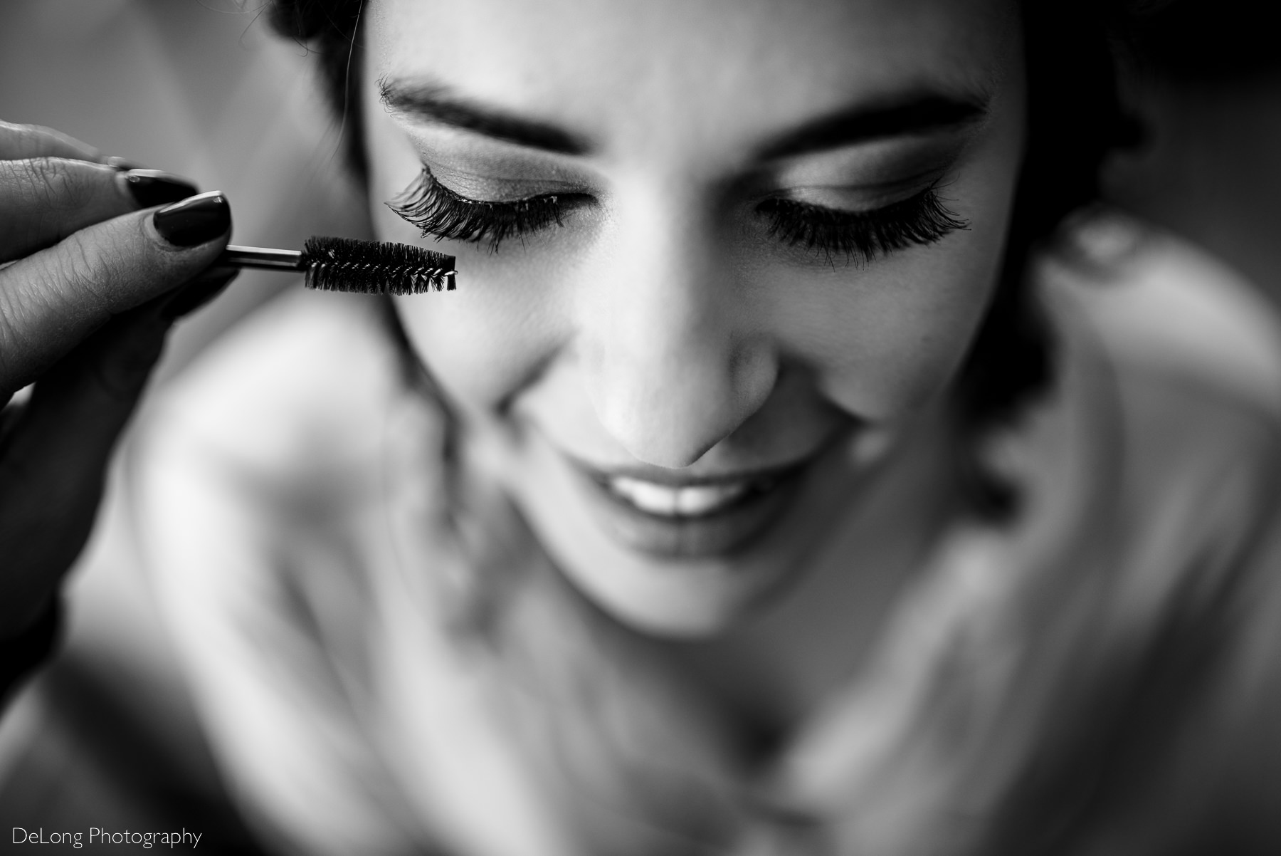 black and white top down photo focused on smiling bride's eyelashes as mascara is applied by Charlotte wedding photographers DeLong Photography