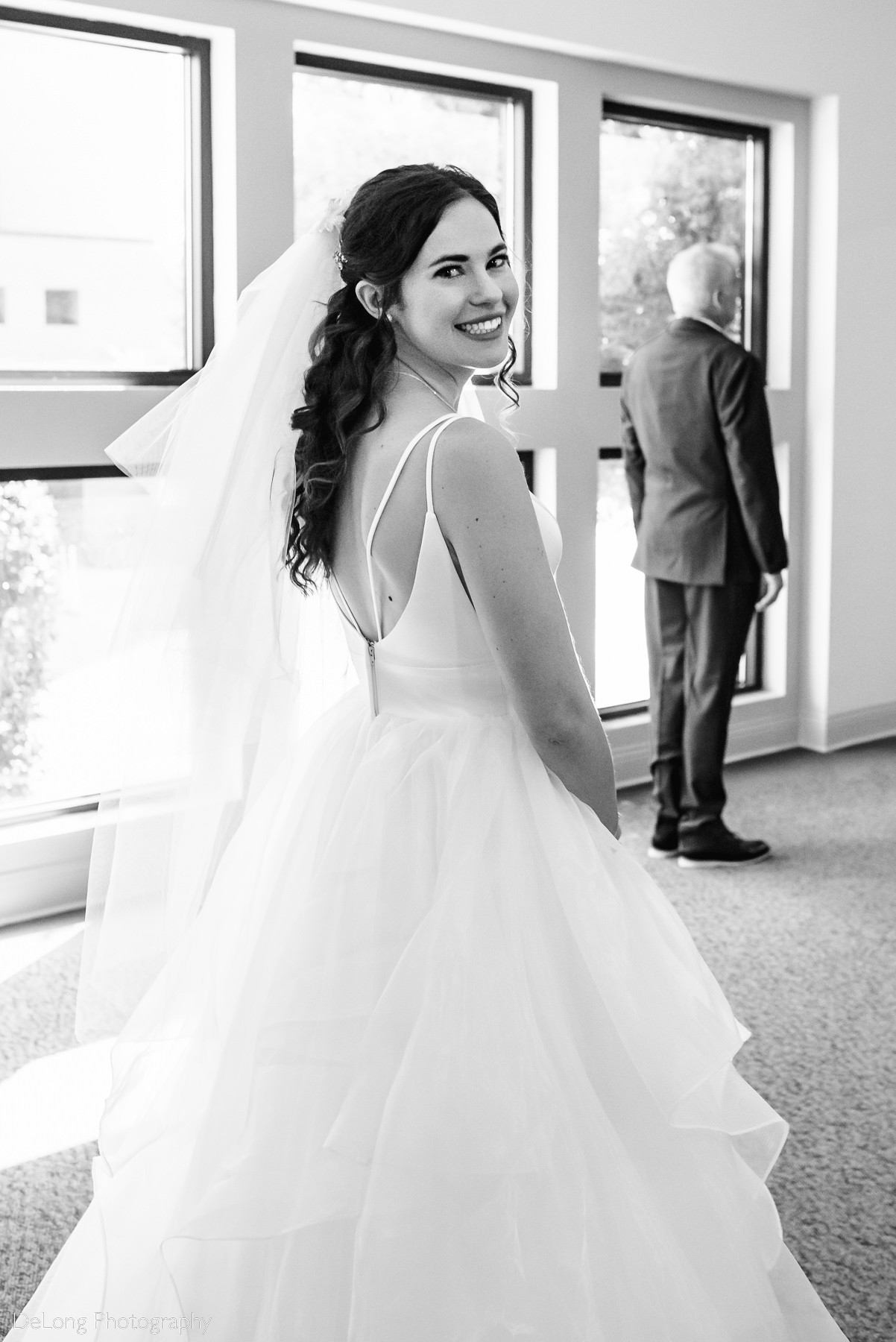 Black and white image of bride smiling looking over her shoulder with her father in the background before a first look by Charlotte wedding photographers DeLong Photography