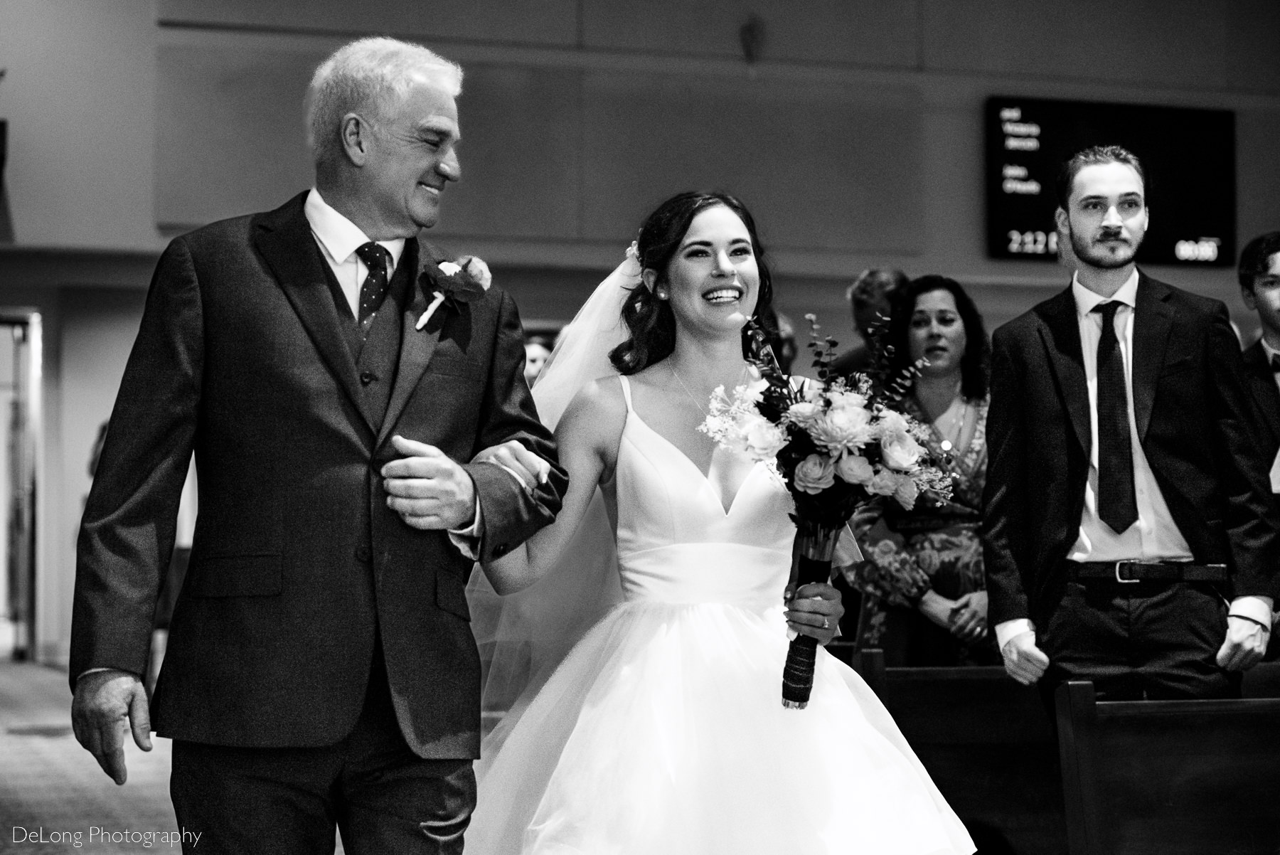 Father of the bride smiling while looking at his daughter who is joyfully smiling at her groom as they walk down the aisle at St Gabriel Catholic Church by Charlotte wedding photographers DeLong Photography