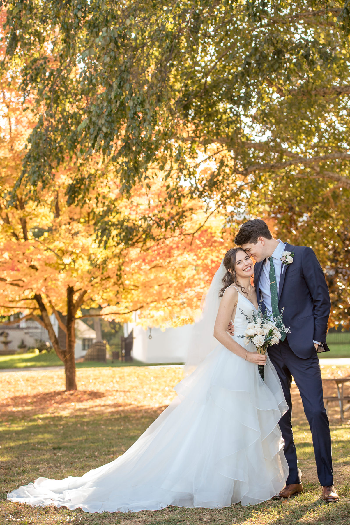 Groom nuzzling into the bride's temple as she smiles at the camera in front of yellow and orange autumn colored trees in Freedom Park by Charlotte wedding photographers DeLong Photography