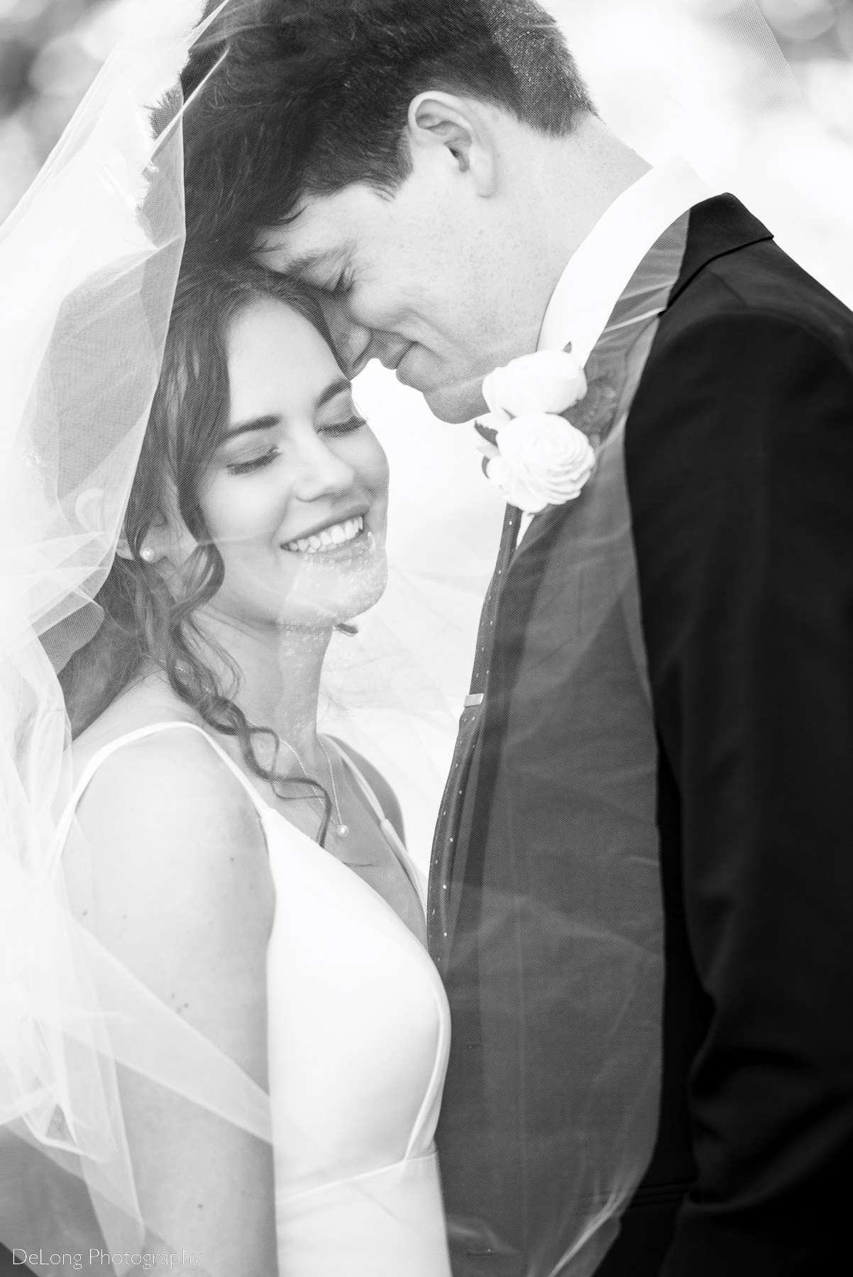 Black and white up-close vertical image of bride and groom under her veil both have their eyes closed smiling as he nuzzles her by Charlotte wedding photographers DeLong Photography